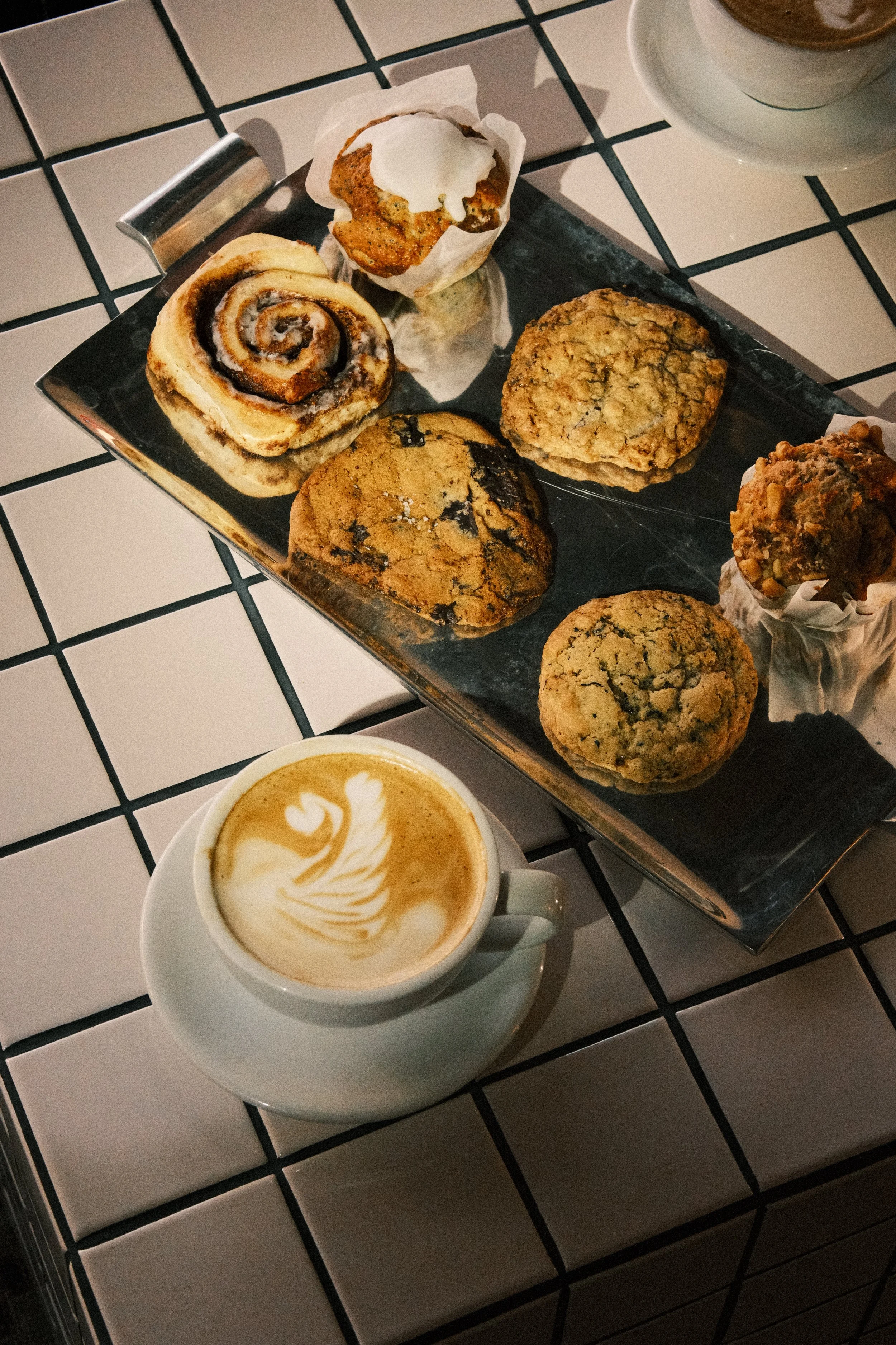 A tray of assorted baked goods including a cinnamon roll, a muffin with white icing, a chocolate chip cookie, and a muffin with a nut topping, alongside a cup of coffee with latte art.