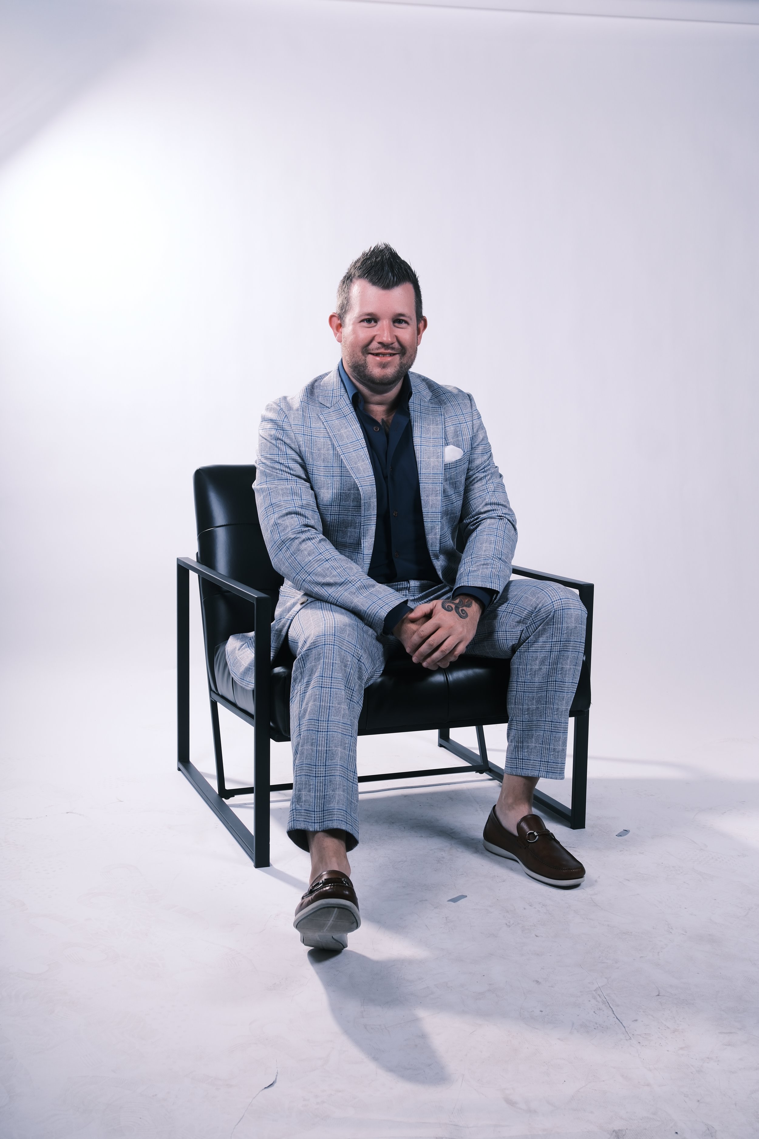 A man sitting in a black chair on a white background, wearing a light gray plaid suit, dark shirt, brown loafers, with tattoos on his left hand, smiling at the camera.