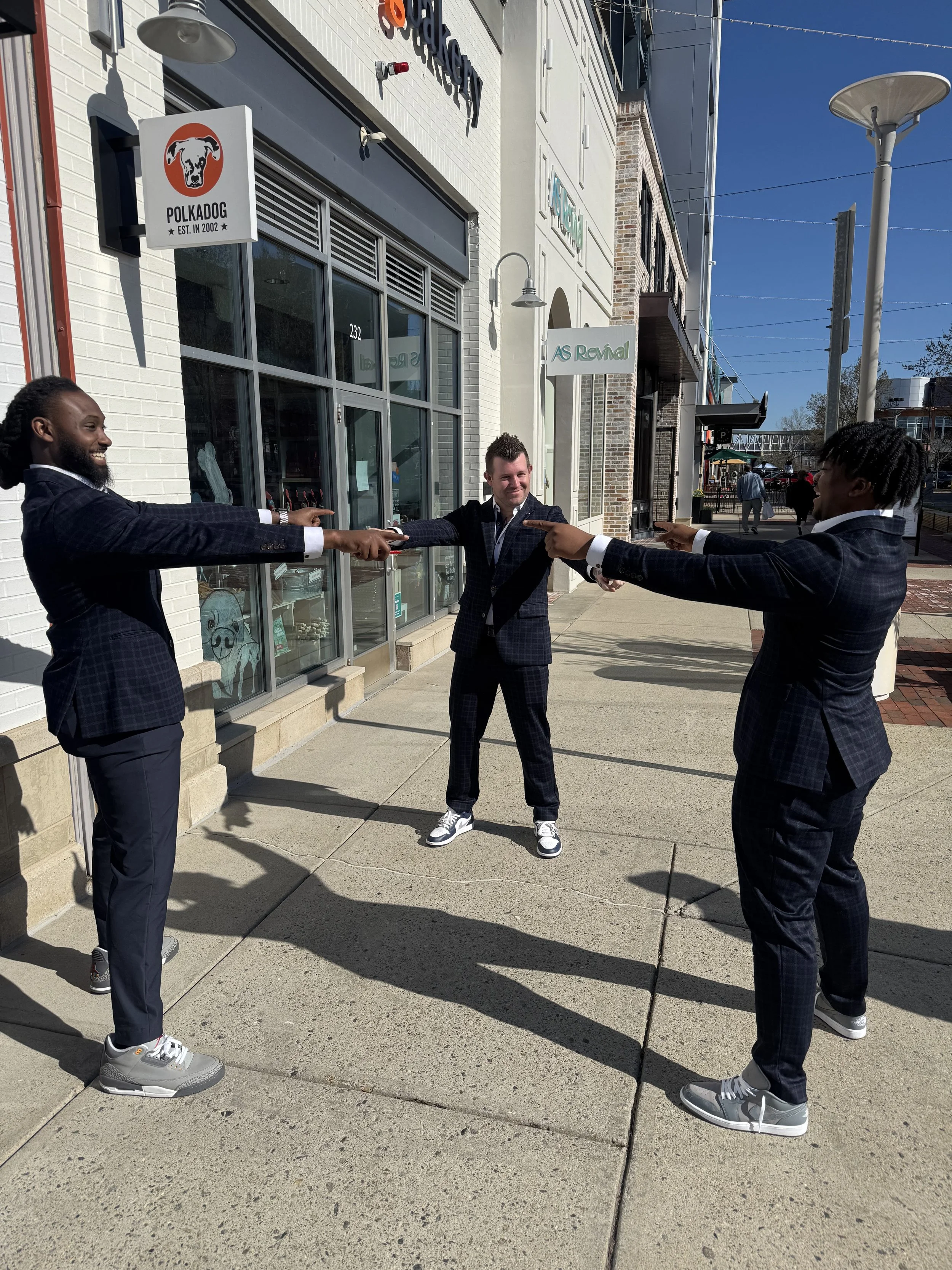Three men standing outside a storefront, all wearing suits, pointing and laughing at each other.