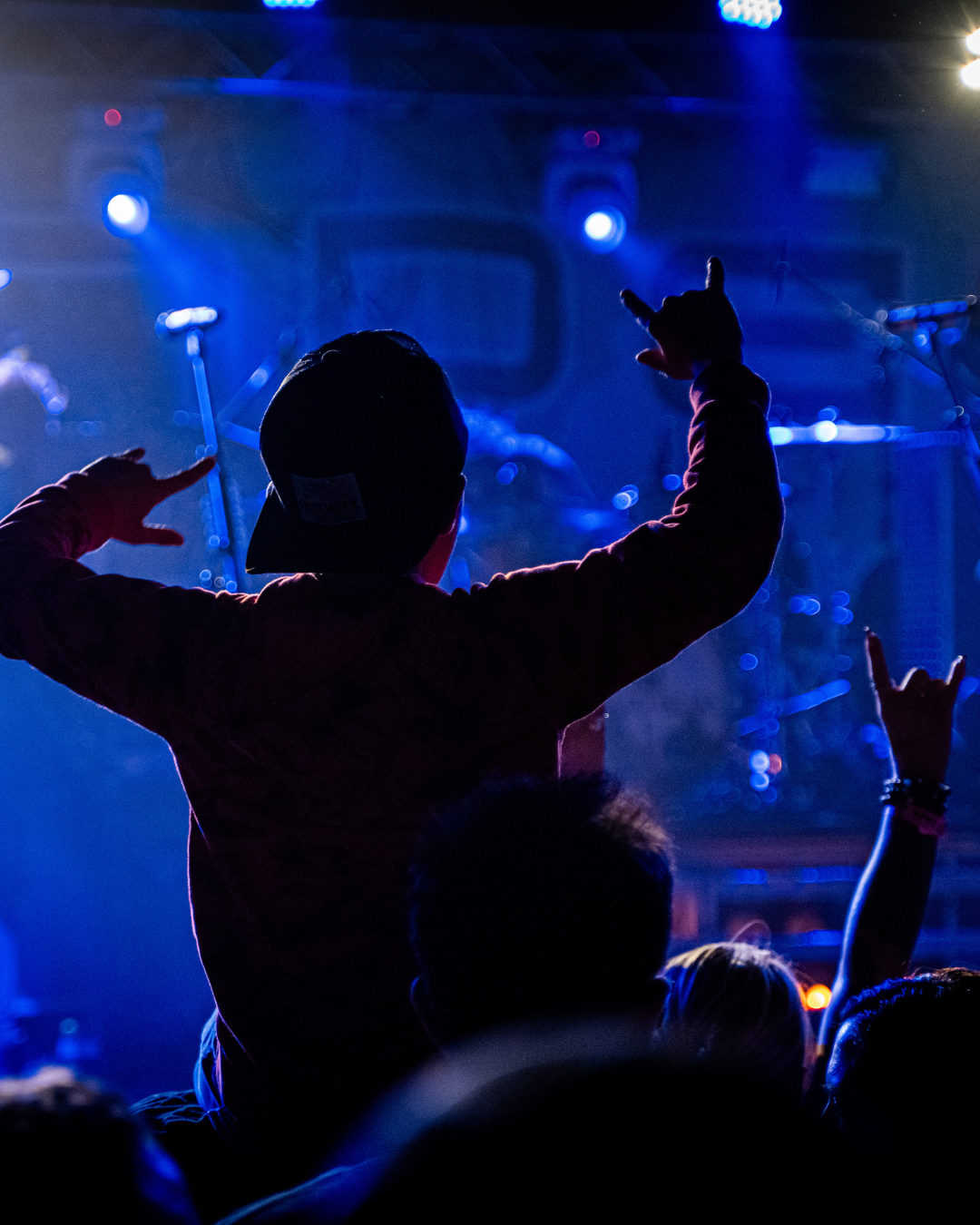 Person at concert raising arms with hands making rock and roll gestures, silhouetted against blue stage lighting.