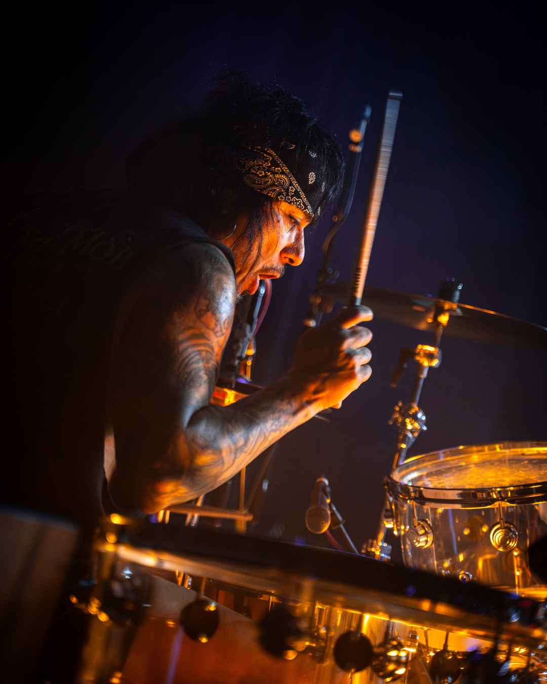 A male drummer with tattoos, wearing a black bandana, performing on stage with drum set illuminated by warm stage lighting.