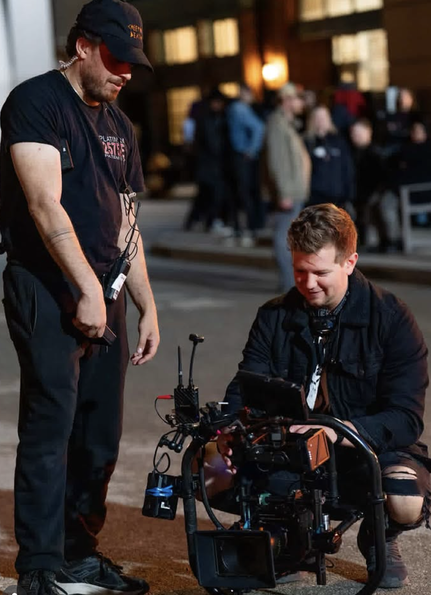 Two men working on a professional camera setup outdoors at night, with a crowd of people in the background.