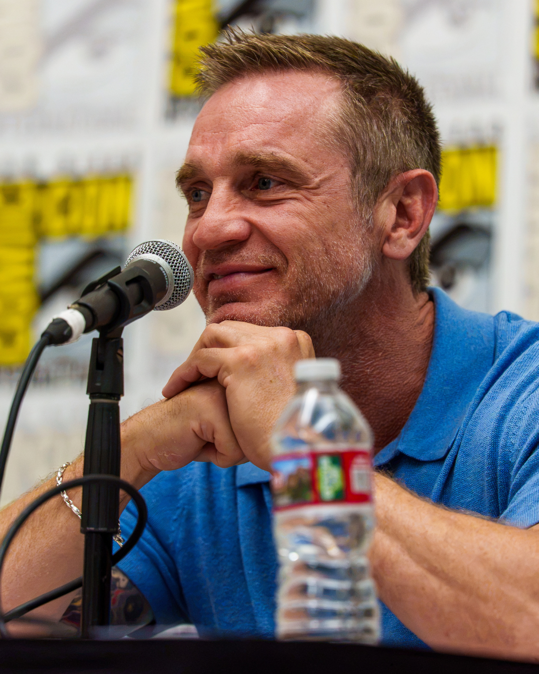 A man with short, styled brown hair, wearing a blue polo shirt, sits at a table with a microphone in front of him. He rests his chin on his folded hands, smiling slightly, with a water bottle in front of him. The background shows blurred posters or signboards.