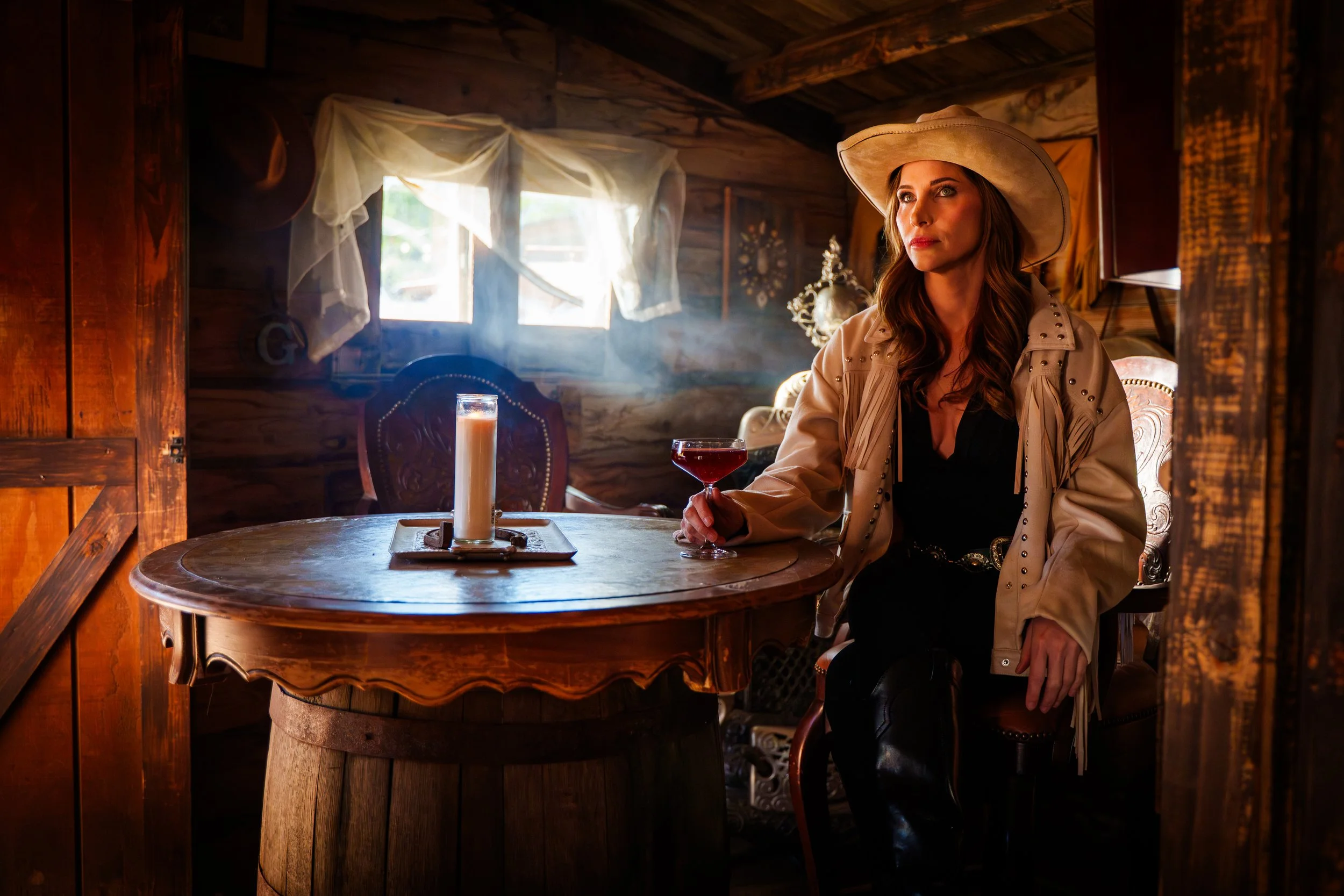 A woman with long brown hair wearing a beige cowboy hat and a beige jacket with stud details, sitting at a round wooden table with a candle and a glass of red drink inside a rustic wooden cabin.