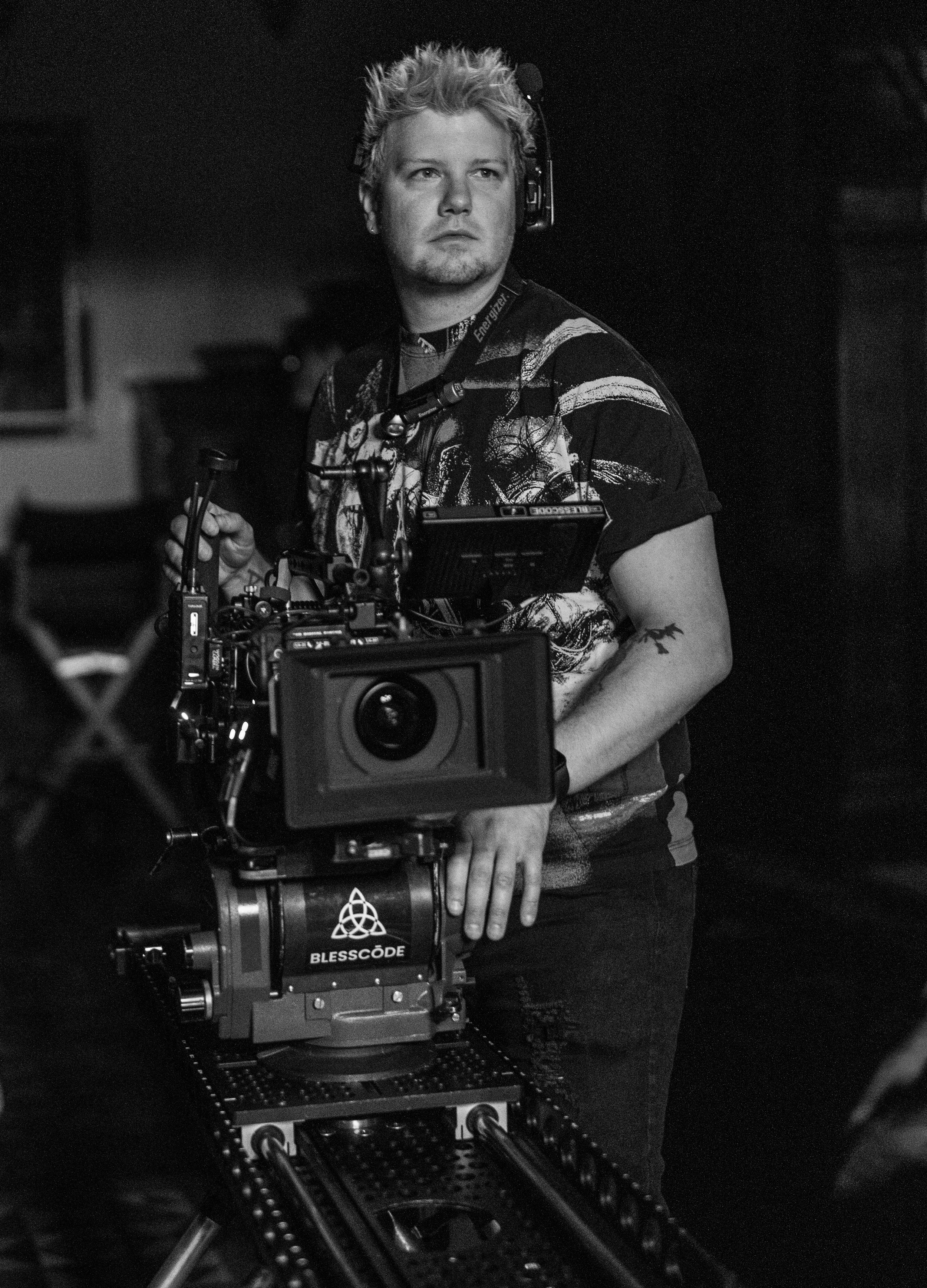 Black and white photo of a man operating a film or camera equipment on a set, wearing headphones, with various electronic devices visible.