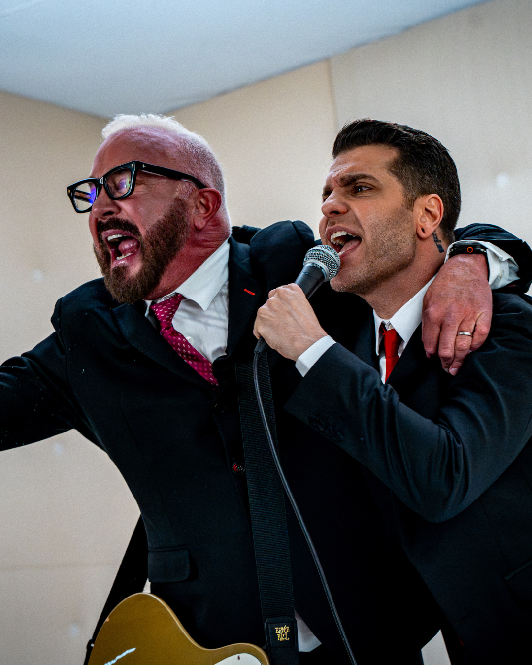 Two men in suits singing passionately, one holding a microphone and the other playing an electric guitar, in an indoor setting.