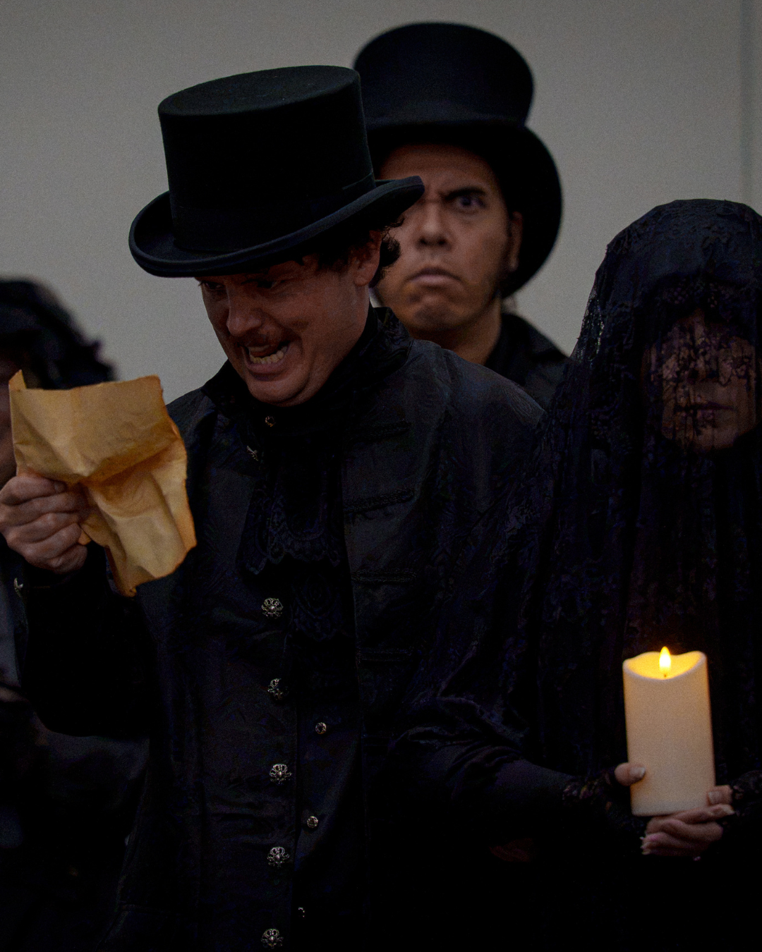 Scene from a theatrical performance featuring actors in dark clothing and top hats, one actor reading from a paper, and others holding candles.