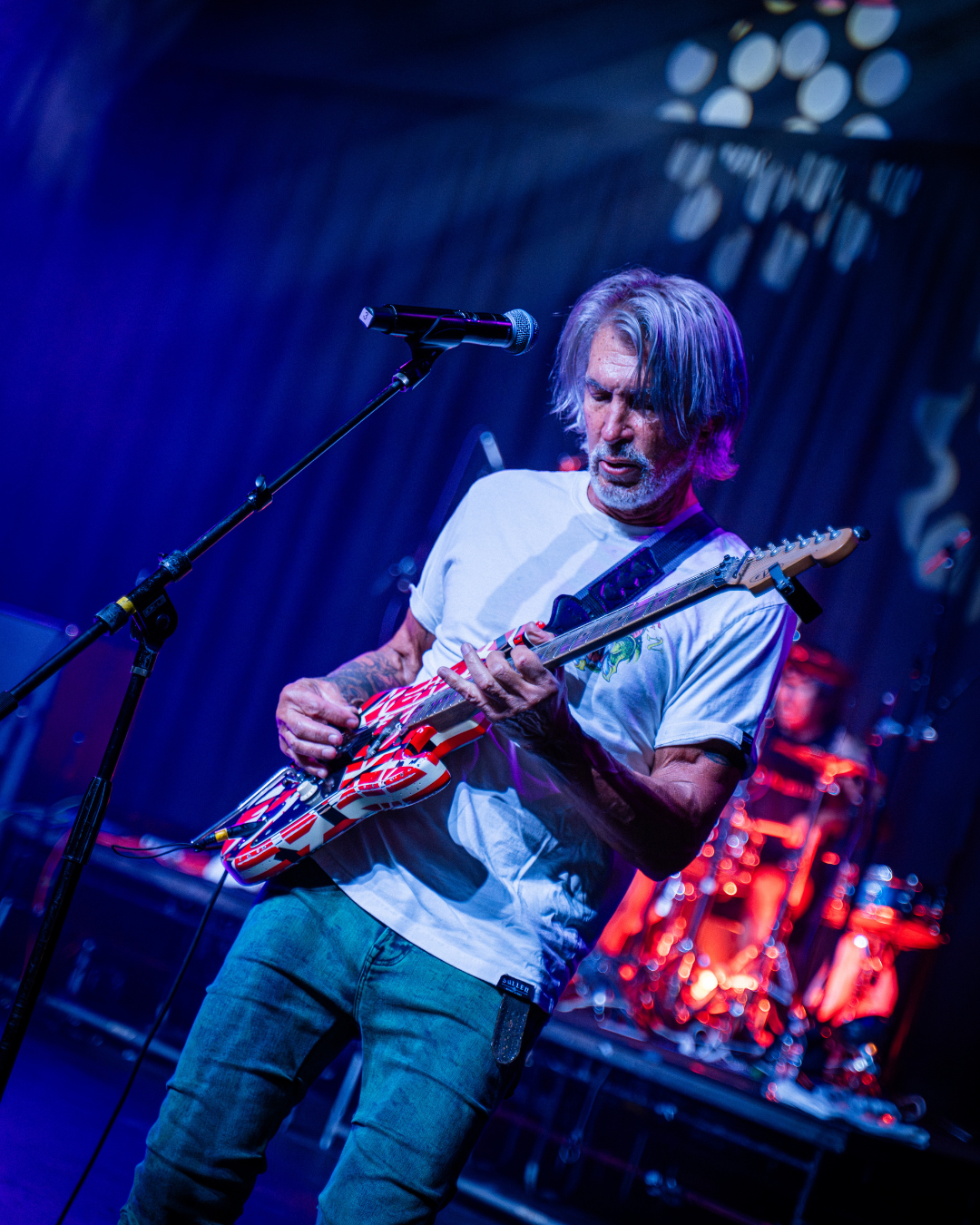 A male musician with gray hair, wearing a white t-shirt, playing an electric guitar on stage with colorful lighting and a drummer in the background.