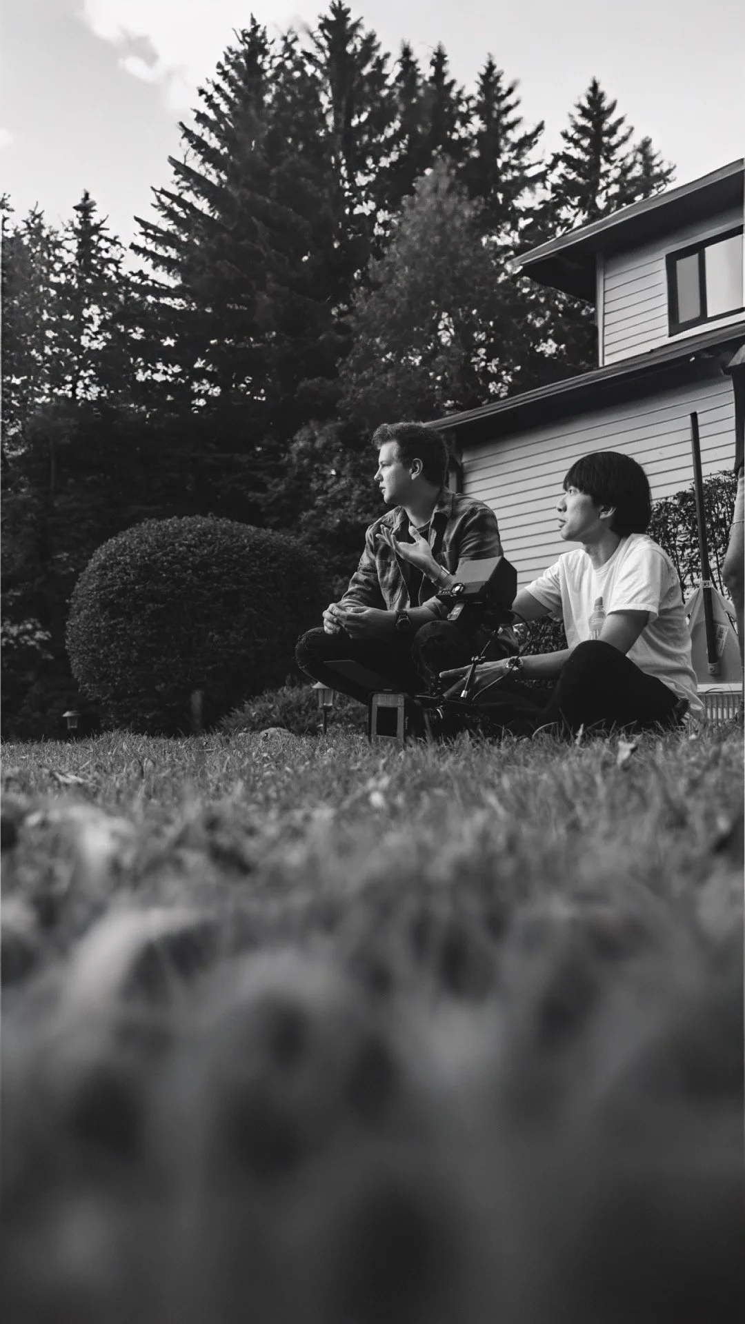 Two people sitting on grass in front of a house, engaging in conversation outdoors, with trees in the background, in black and white.