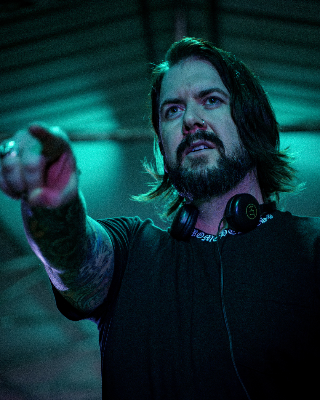 A man with long dark hair, a beard, and tattoos on his arm, wearing headphones around his neck, pointing or reaching out with his finger.