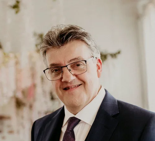 A man in a suit and tie with glasses smiling in an indoor setting with a blurred background.