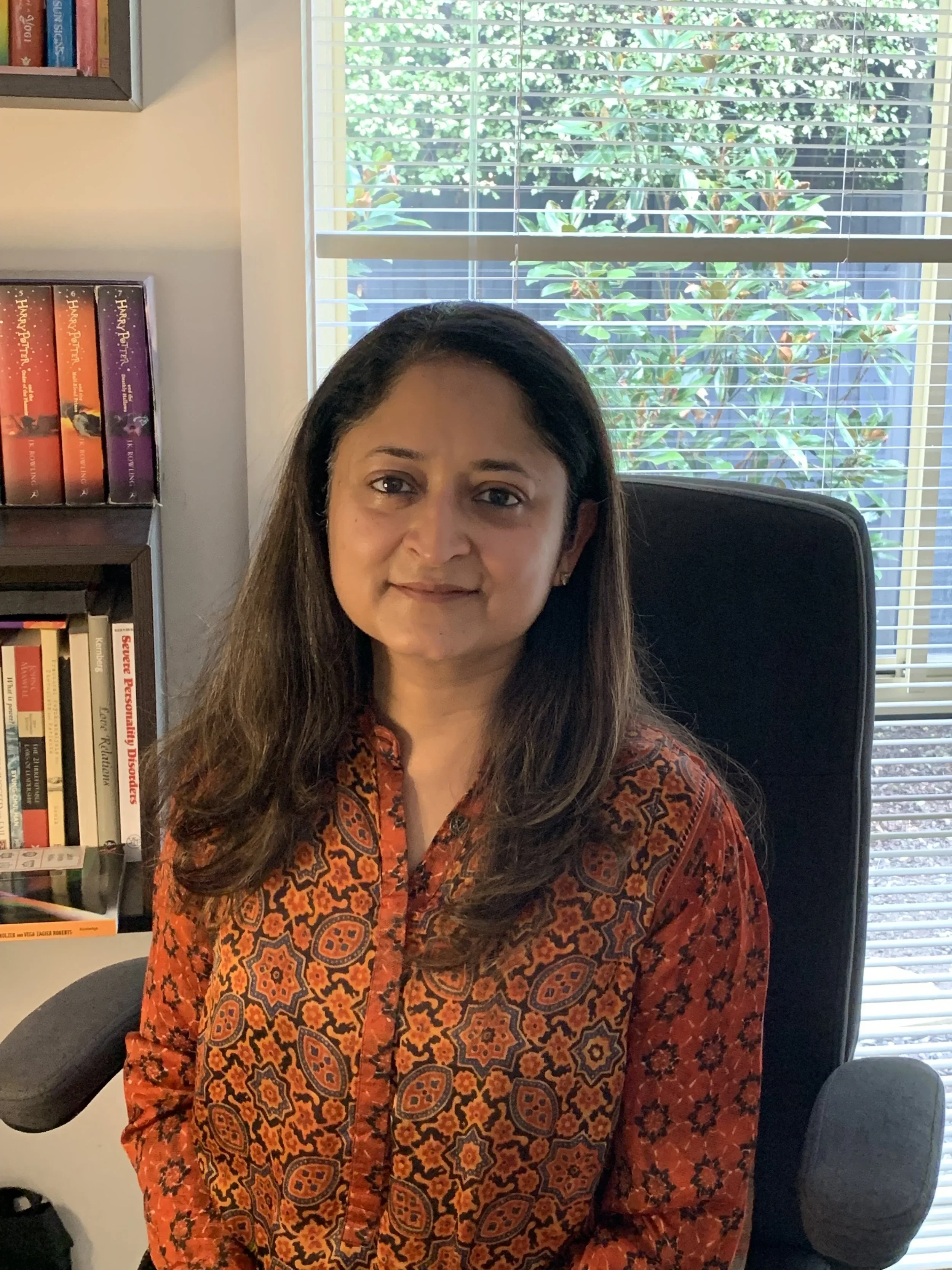 A woman with long dark hair and a patterned orange and red blouse sitting in a black office chair, in a room with a window and a bookshelf filled with books in the background.