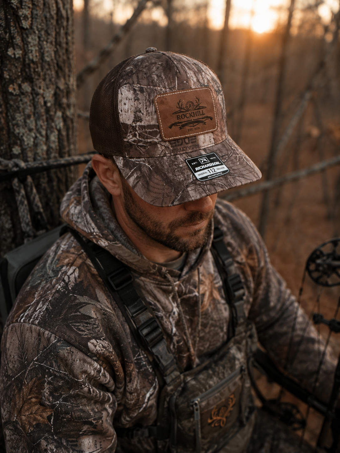 A man in camouflage outdoor gear, wearing a camouflage cap with a "Rockhill" logo, is sitting on a tree in a forest during sunset, with a bow and arrow visible.