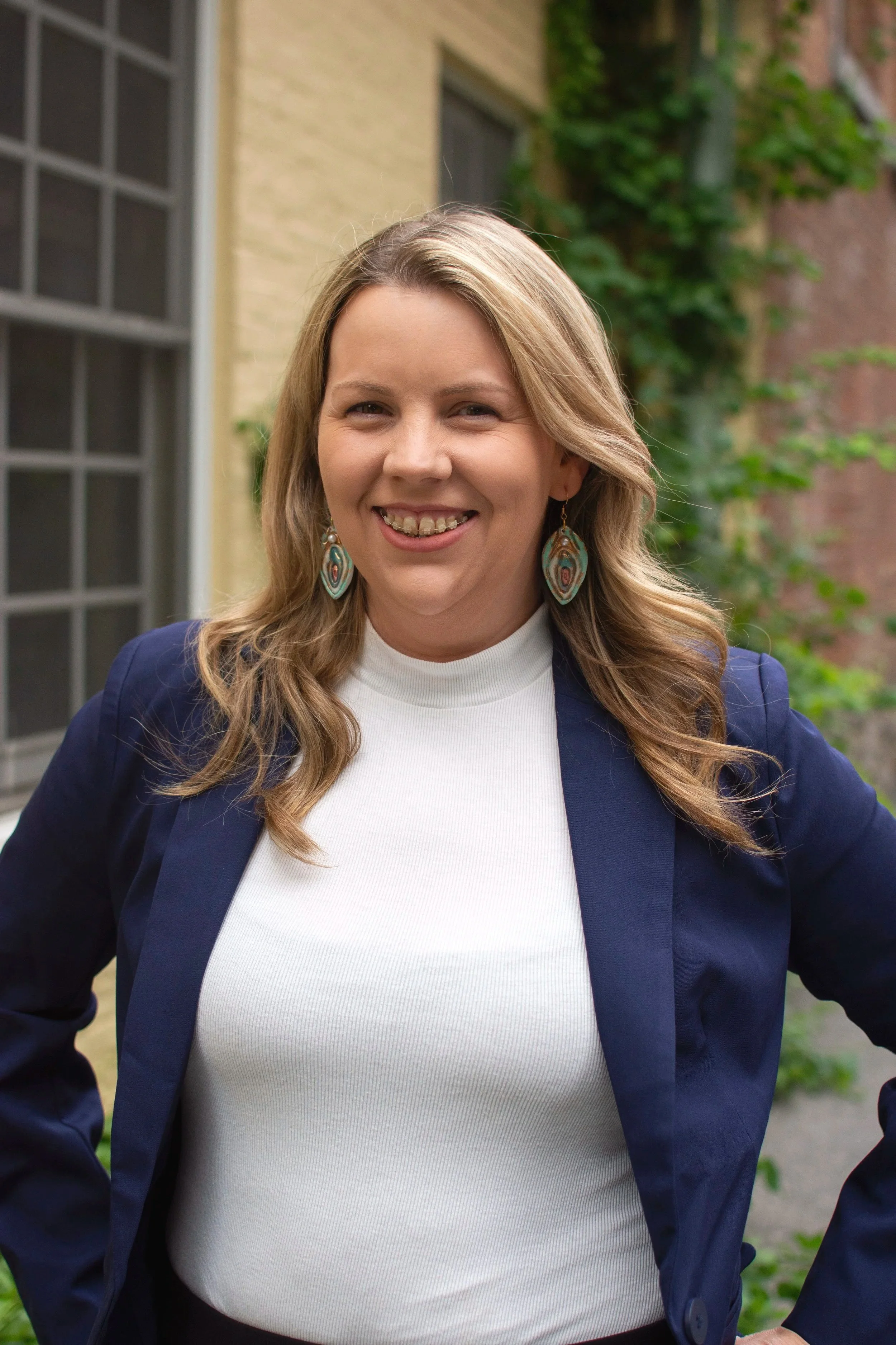 Carissa O'Rourke, wearing a navy blazer and large turquoise earrings, standing outdoors in front of a building with yellow brick and a window, with greenery in the background.