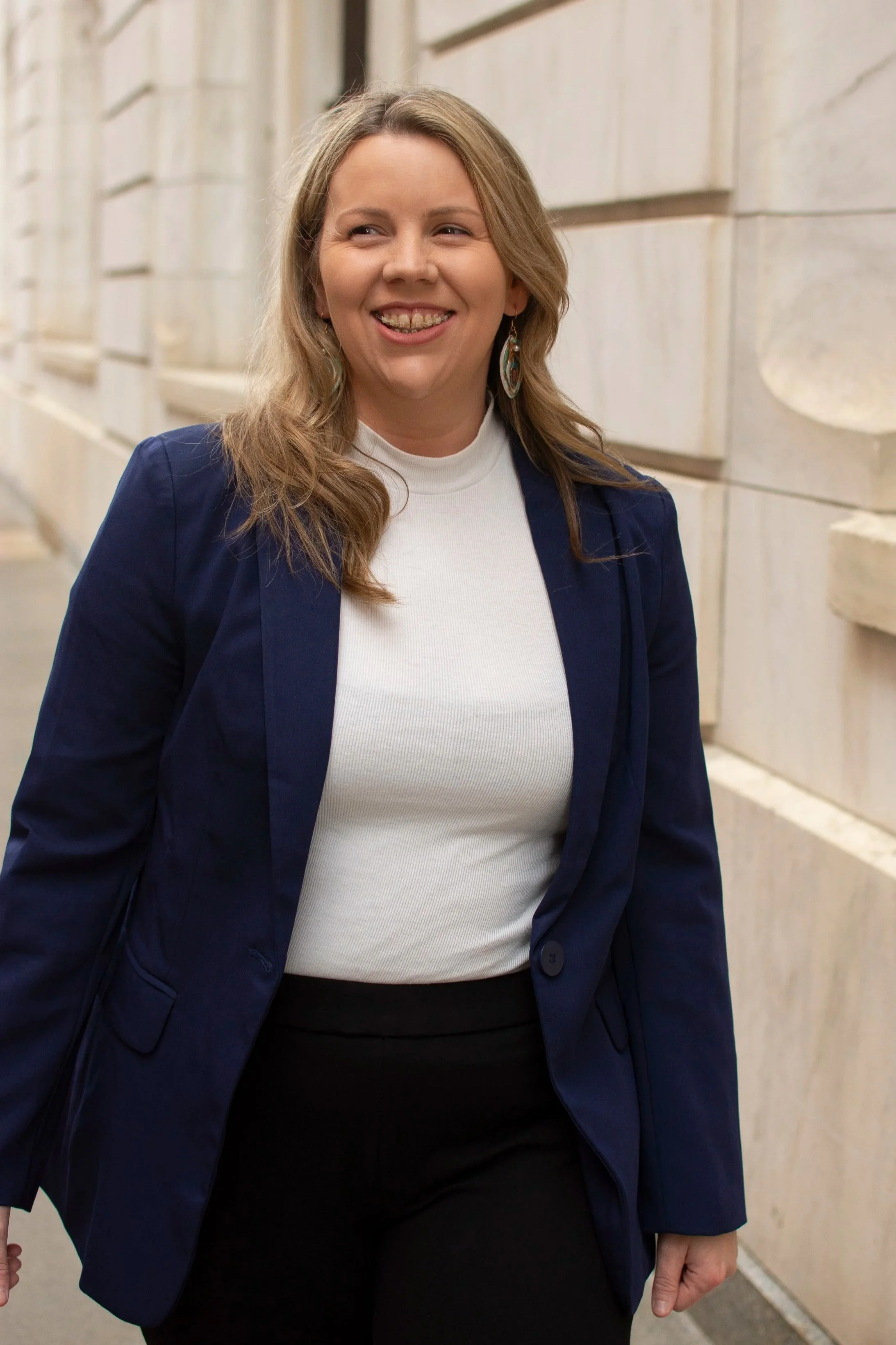 Carissa O'Rourke with long, wavy blonde hair wearing earrings, a white turtleneck, a navy blazer, and black pants, standing against a stone building wall.