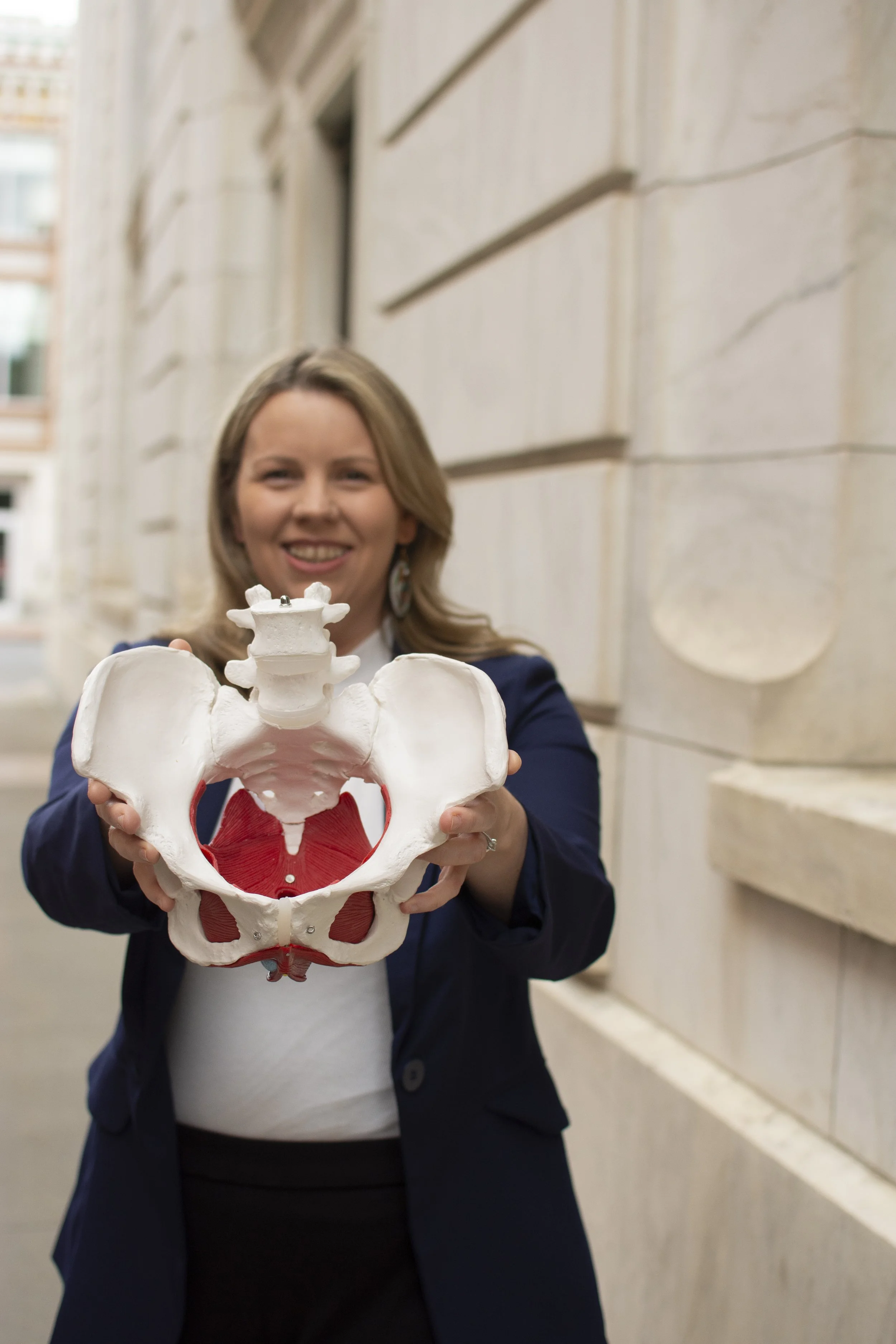 Carissa O'Rourke smiling and holding a model of a human pelvis and lower spine, standing outdoors in front of a stone building.