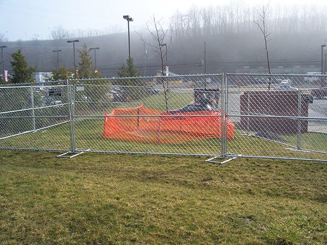 Construction fence with orange safety barrier and a piece of heavy equipment behind it in a grassy area near a parking lot with trees and foggy conditions in the background.