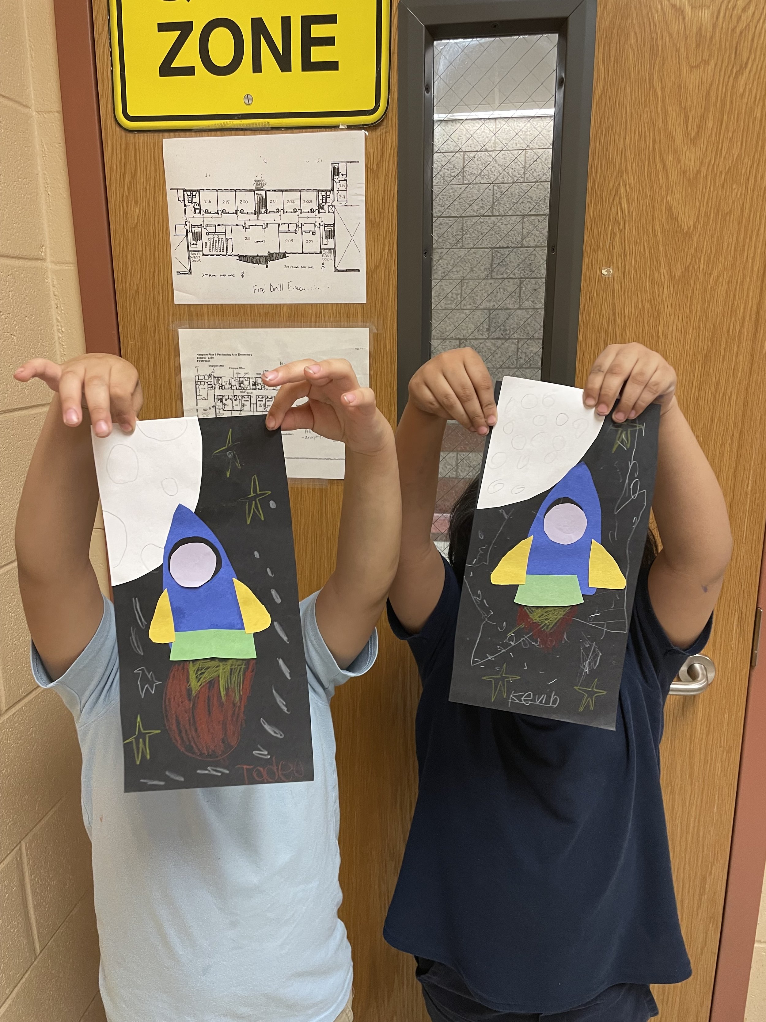 Children holding up paper cutouts of rockets in front of a door with a school map and a yellow zone sign.