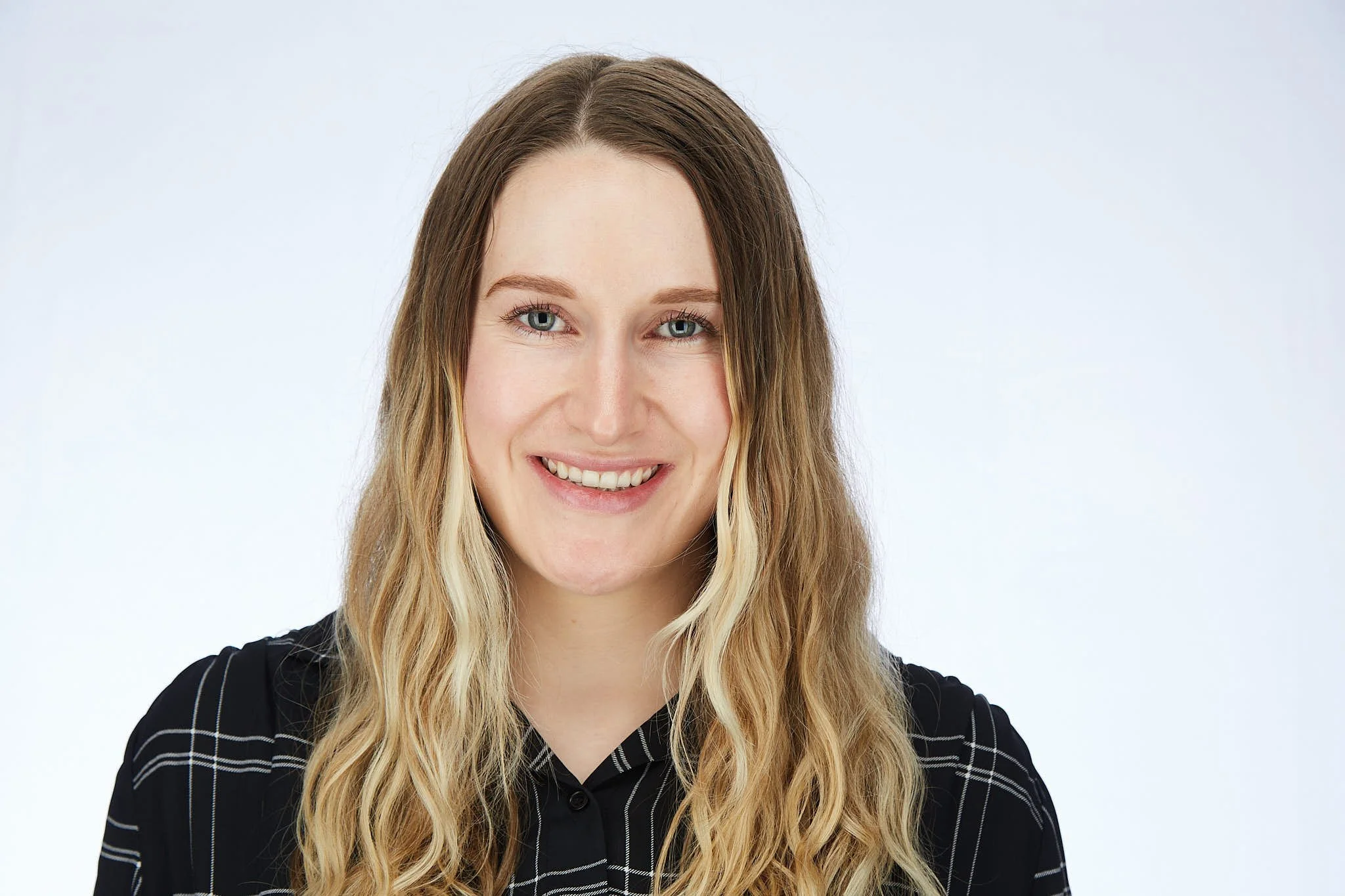 Portrait of a woman with long wavy blonde hair, blue eyes, wearing a black top with white pinstripes, smiling against a plain light background.
