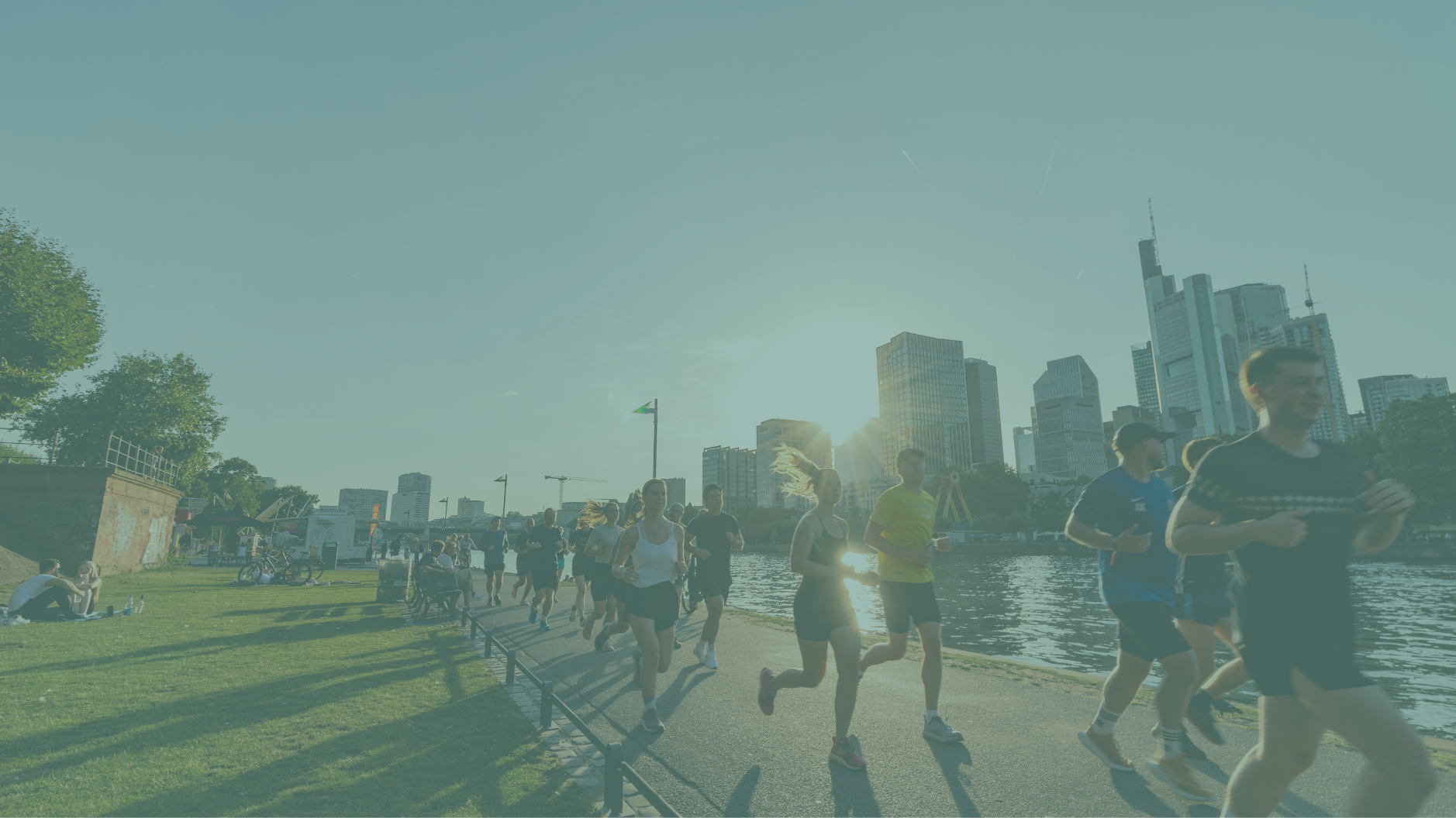 People jogging along a riverfront promenade in an urban park during sunset, with city skyscrapers in the background.