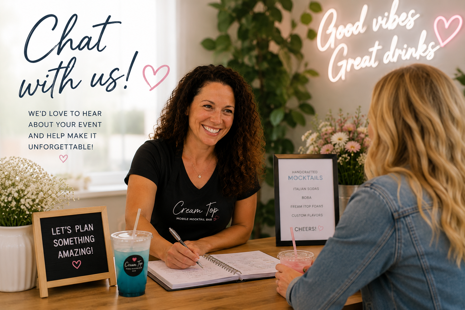 A woman with curly hair smiling and taking notes at a mobile mocktail bar counter, talking to a customer with long blonde hair, seated across from her. The setting is bright and welcoming with signs that say "Chat with us!" and "Good vibes, Great drinks." There are flowers and decorative signs on the counter promoting handcrafted mocktails and encouraging planning for something amazing.
