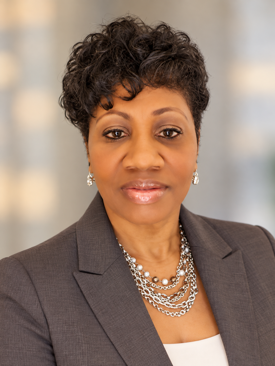 Professional woman with short curly black hair wearing a gray blazer, white top, pearl and silver jewelry, and earrings, posing in front of a blurred neutral background.