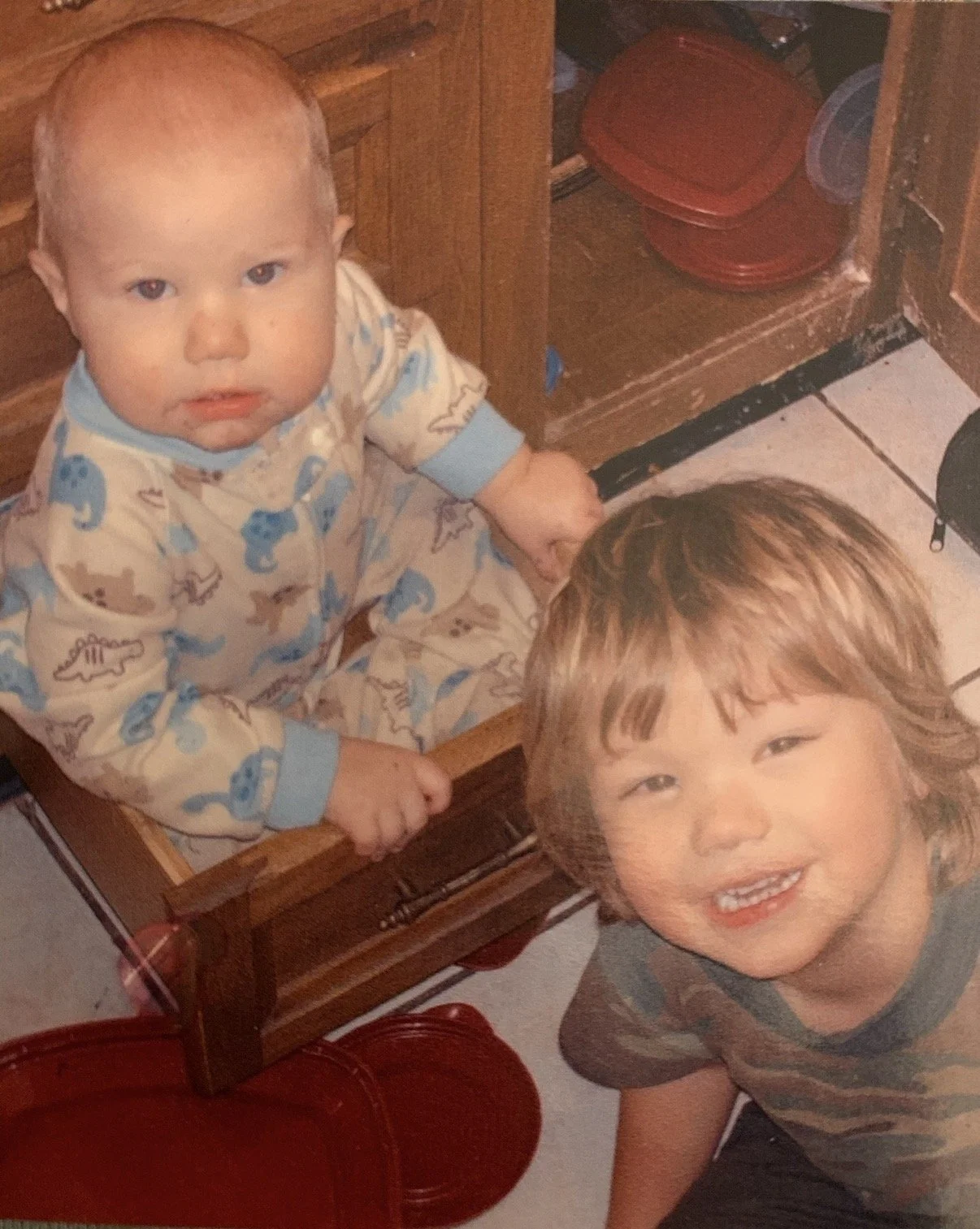 Two young children in a kitchen; one child in pajamas with blue and tan dinosaur prints sitting on a wooden chair, looking up at the camera, and the other child with light brown hair, smiling and looking up at the camera. Kitchen cabinets and a red dish rack are visible in the background.