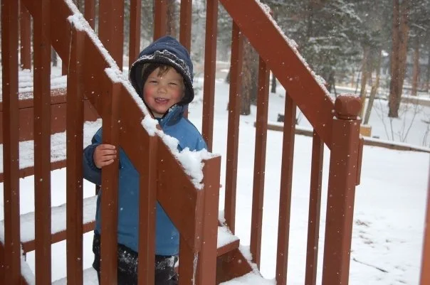 A young boy wearing a blue winter jacket and a dark hat is smiling while playing in the snow behind a wooden staircase railing.