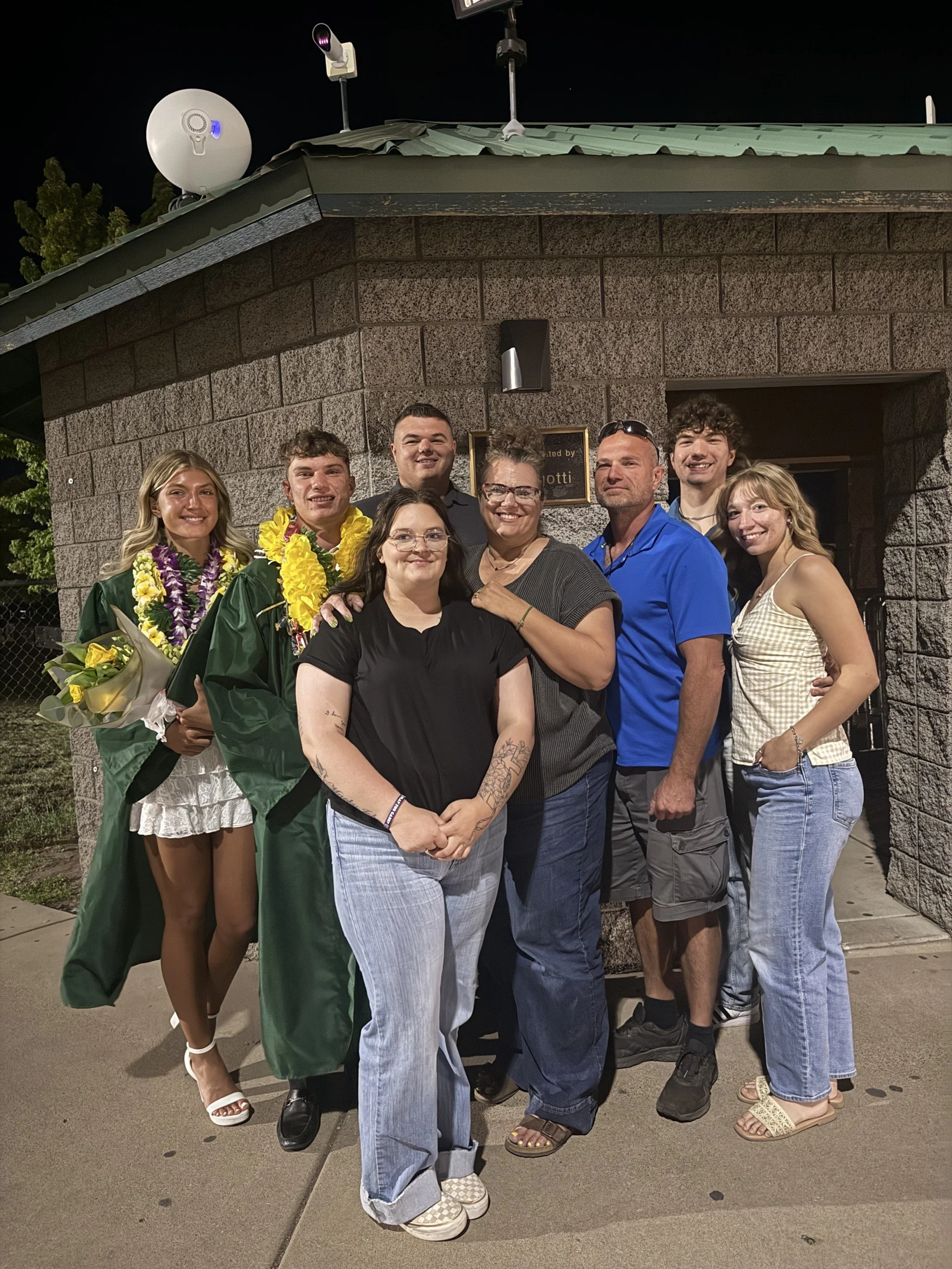 A group of eight people, including a graduate in a green gown and cap, posing for a picture outside a building at night. The group includes men and women of varying ages, some wearing leis, and they are smiling and standing close together.