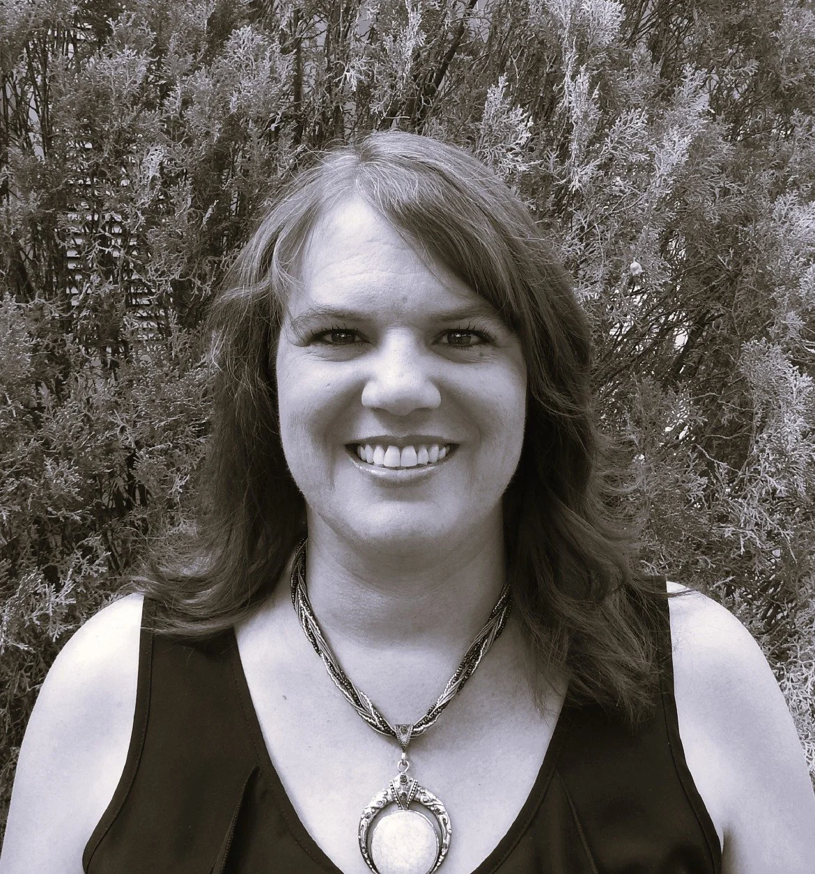 A woman with shoulder-length hair smiling in front of a bush with small leaves.