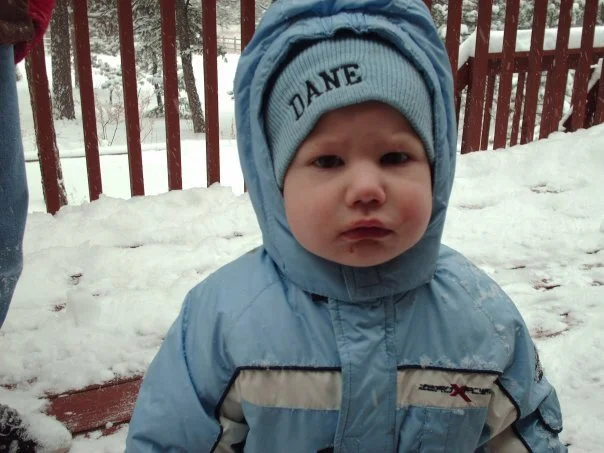 Child in a blue winter coat and gray beanie hat with the name 'DANE' on it standing outdoors in snow, with a red fence in the background.