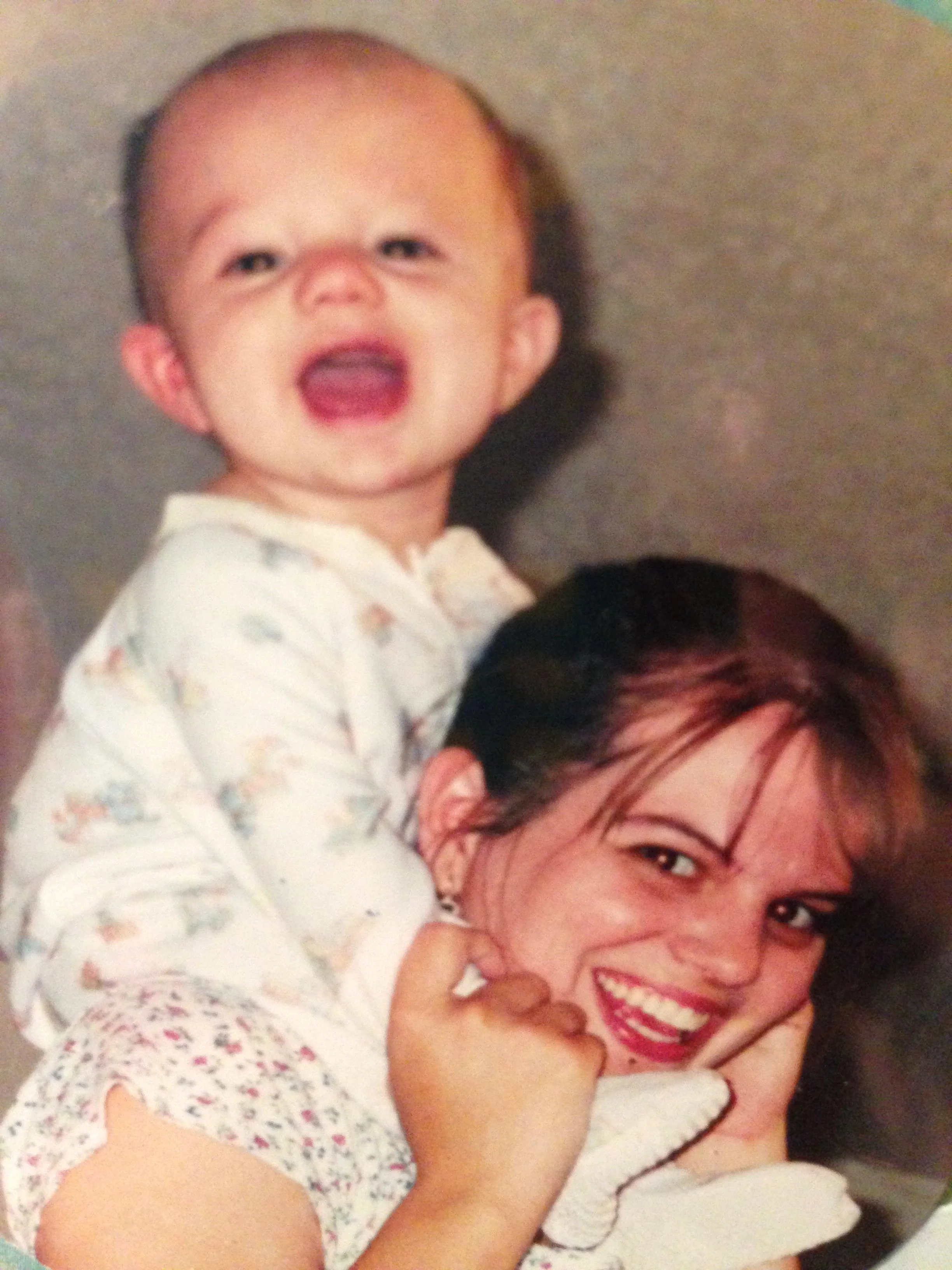 A woman with dark hair and red lipstick smiling joyfully, holding a young child with short hair, light skin, and wearing a white shirt with colorful patterns, lying on her shoulder.