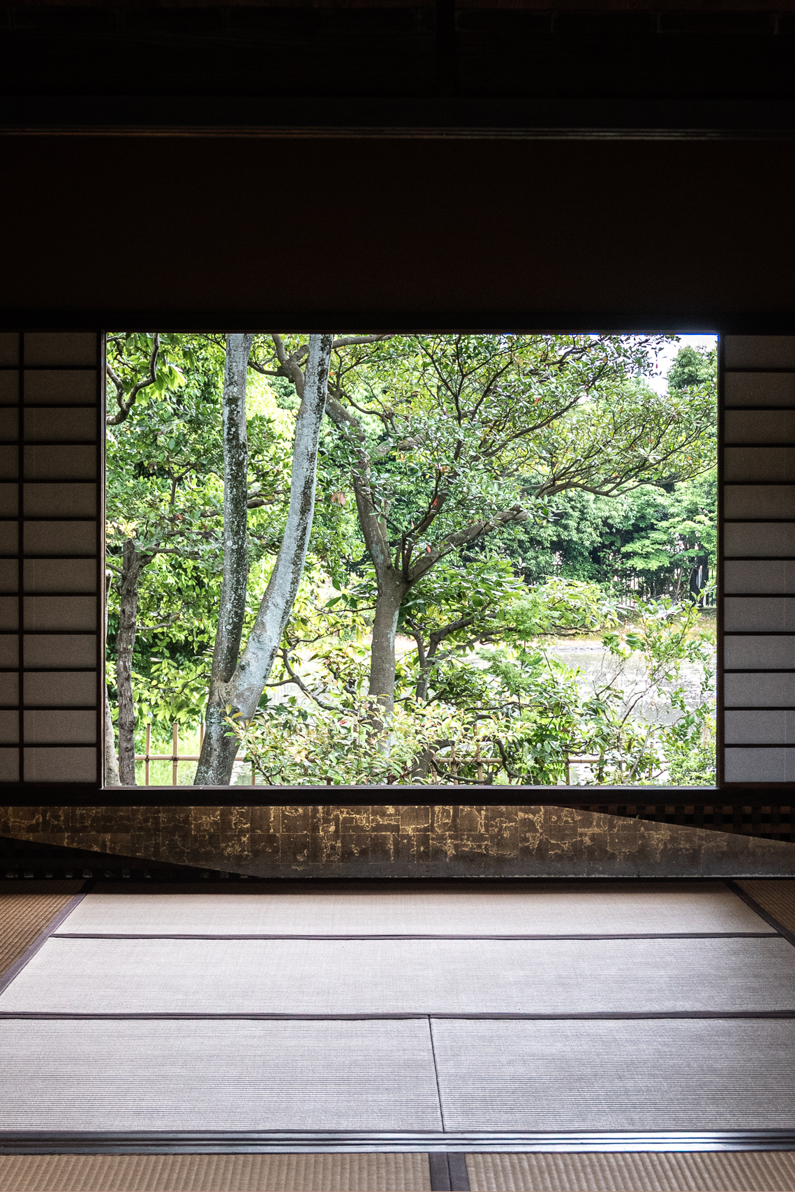 View from inside a traditional Japanese room with tatami mats, looking out through a rectangular window at lush green trees and a body of water in the background.
