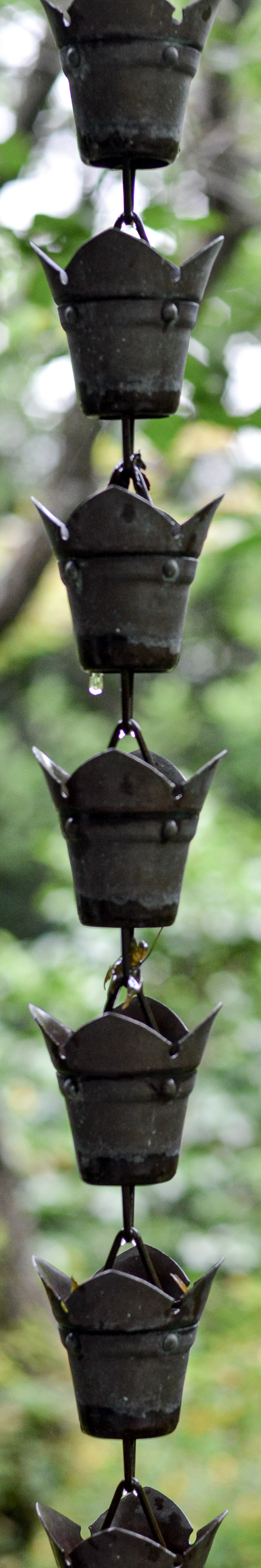 Multiple metal crown-shaped ornaments hanging on a chain with water droplets, outdoors with blurred greenery in the background.