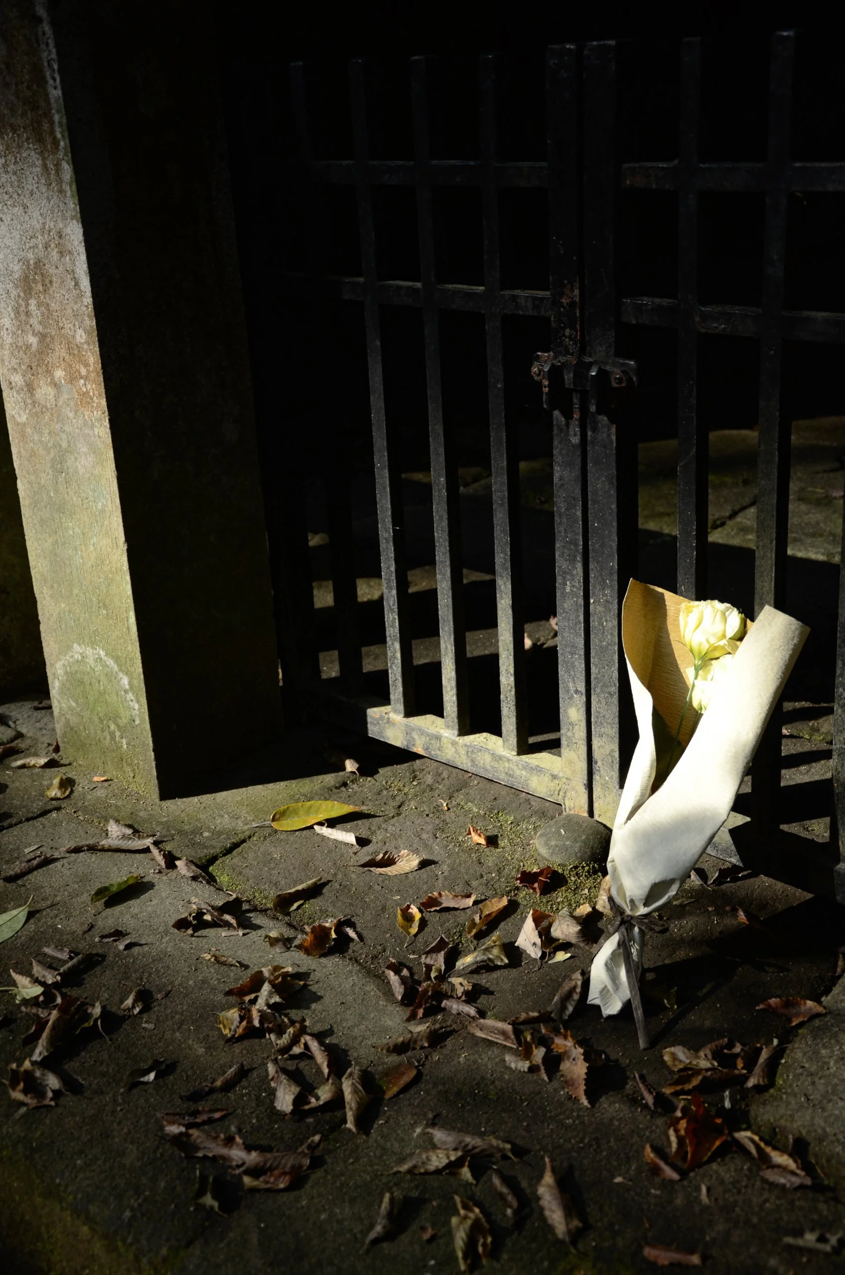 A black metal gate with a padlock, set in a concrete wall, with a bouquet of white flowers wrapped in paper leaning against it, surrounded by dried leaves on a stone pavement at night.