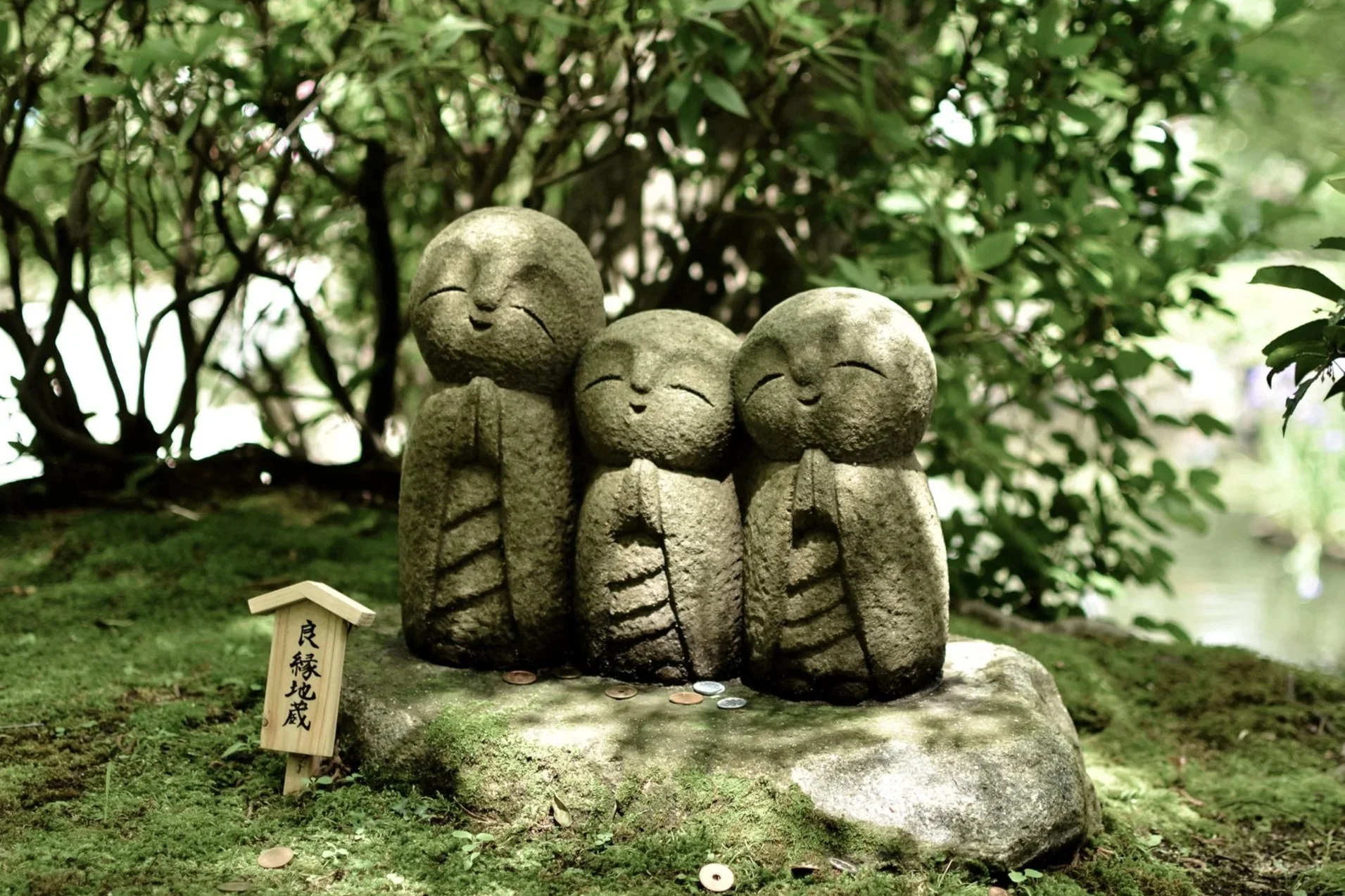 Three stone statues of smiling monks in a lush green garden, with coins at their feet and wooden plaque with Japanese writing nearby.