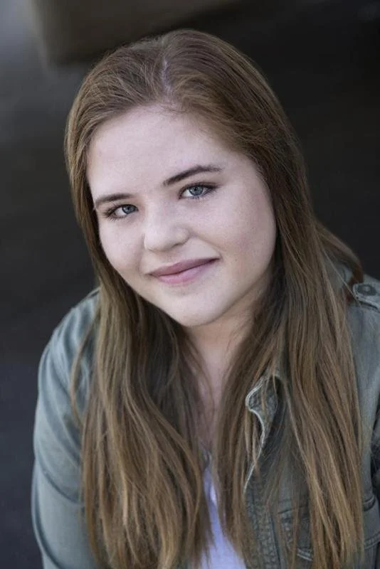 Portrait of a young woman with long, light brown hair and blue eyes, smiling at the camera, wearing a light-colored jacket.