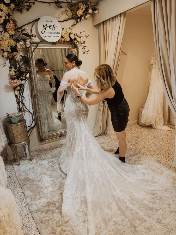 A bride trying on a wedding dress with a helper adjusting the back in a fitting room decorated with flowers and a large mirror.