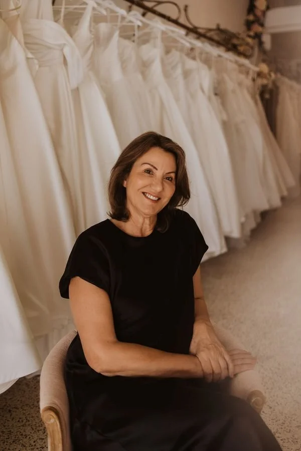 Connie Di Gennie  in a black dress sitting on a chair, smiling, with white wedding dresses hanging behind her. She is in the Showroom of Accapella Bridal in Wollongong NSW.