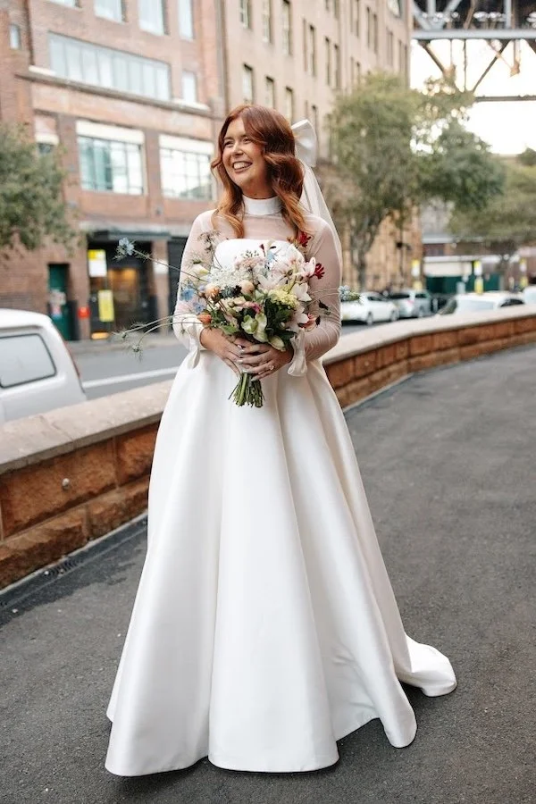 A smiling woman in a white wedding dress holding a bouquet of flowers on a city street. She is a satisfied bride who bought her dress at Accapella Bridal