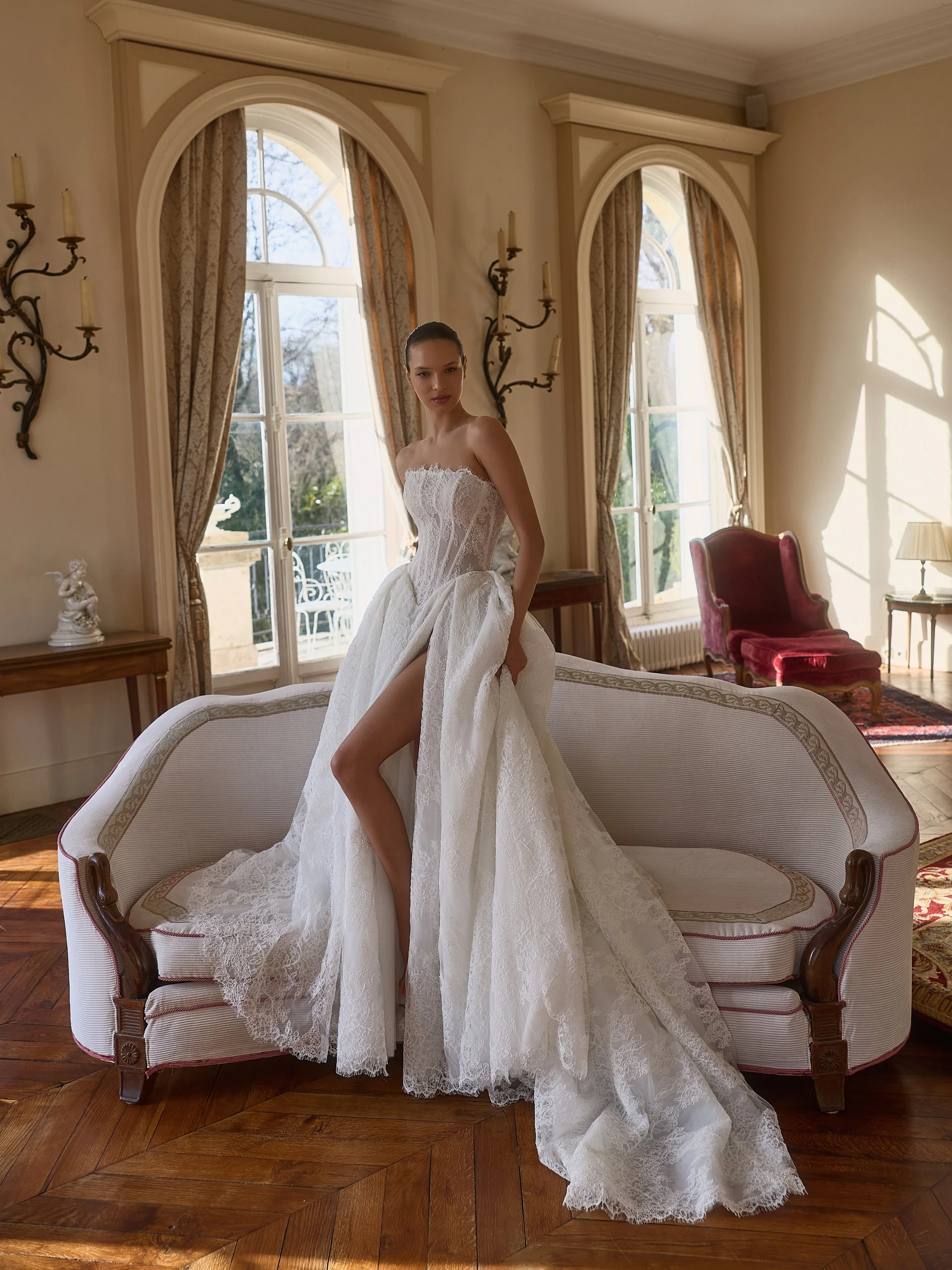 Bride in a strapless lace wedding dress by Maison Yana posing on an upholstered sofa in an elegant sunlit room