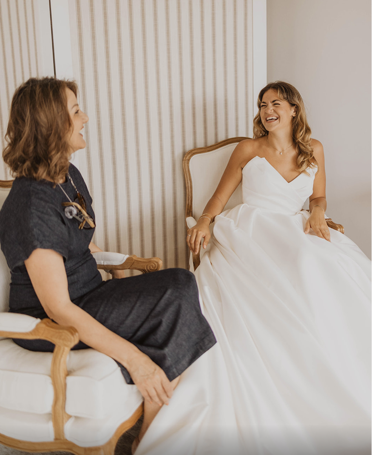 Two women sitting and laughing together in a room; one in a wedding dress, the other in a dark dress.