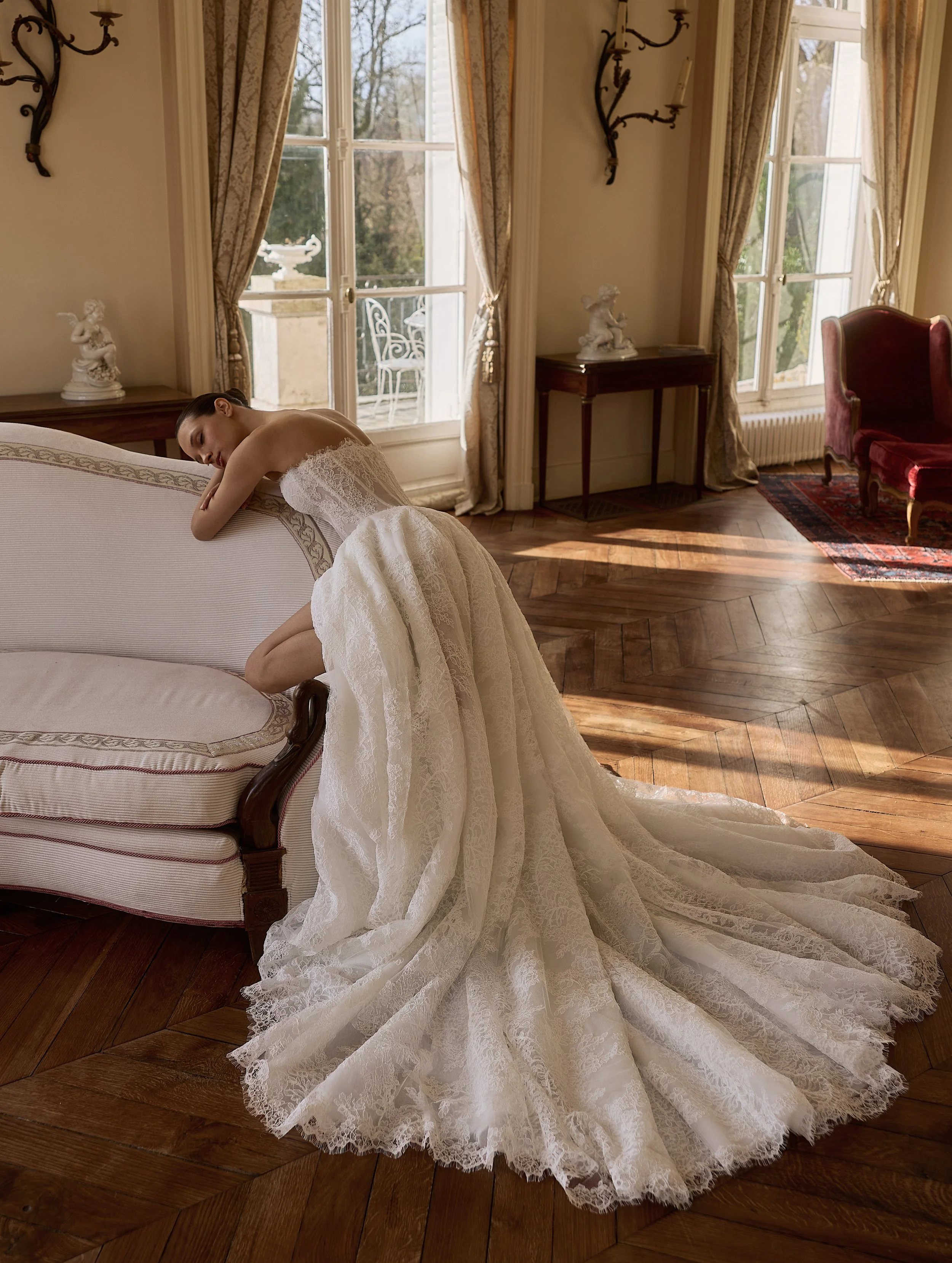Bride in a white lace wedding dress by Maison Yana reclining on an ornate sofa in an elegant room with tall windows, curtains, and antique furnishings.