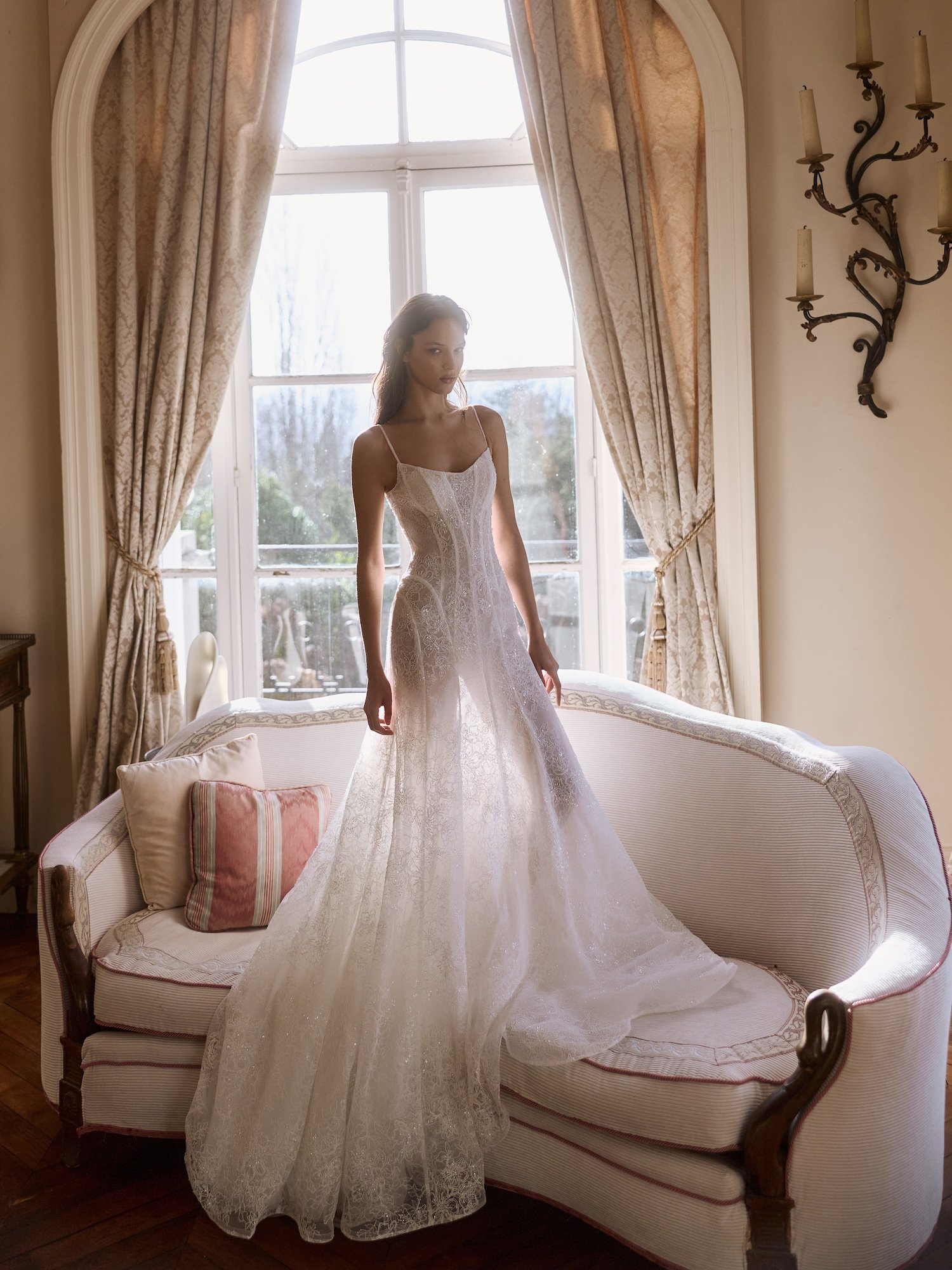 Woman modeling a white lace wedding dress by Maison Yana in an elegant room with a large window, curtains, and vintage sofa