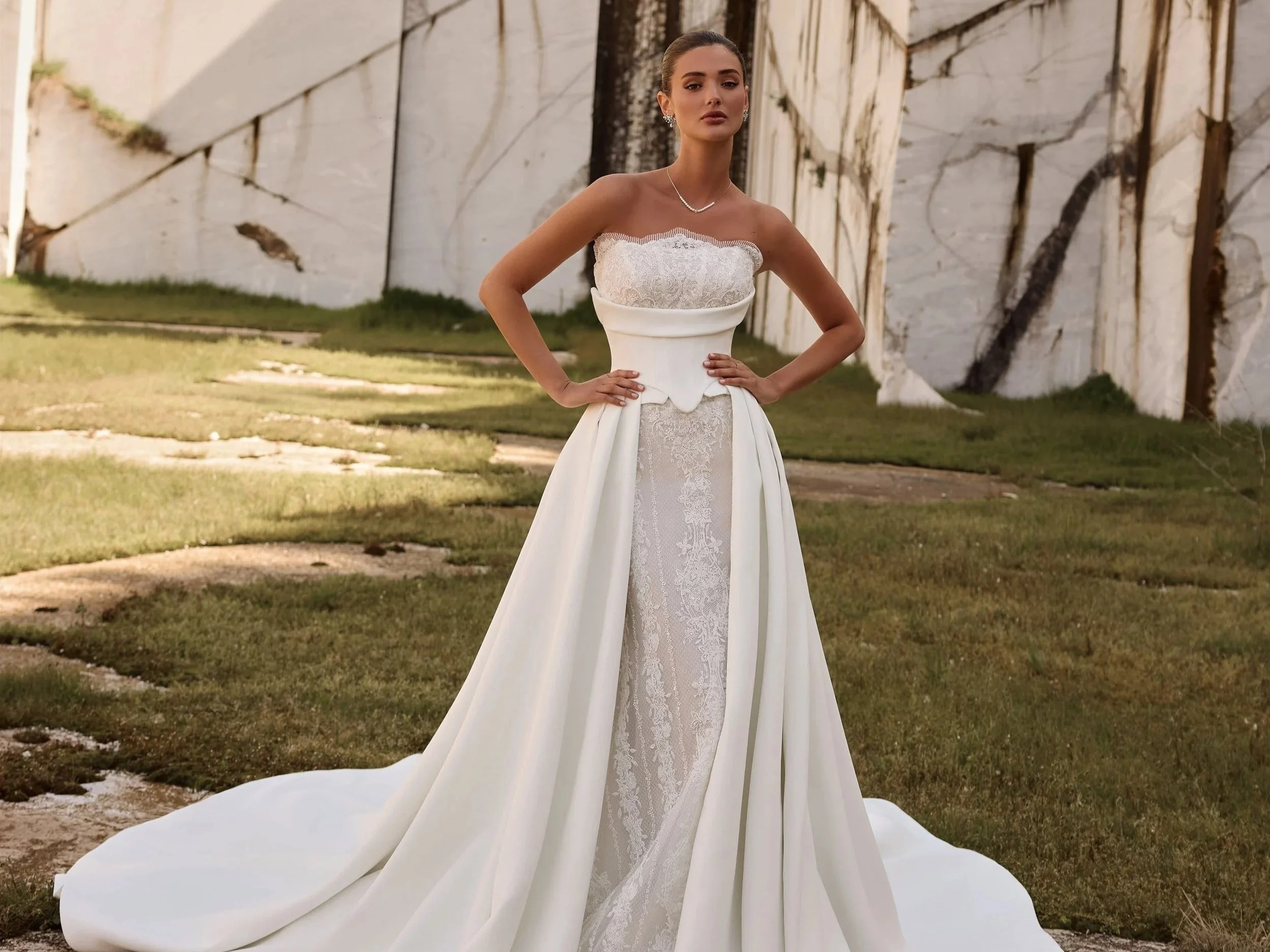 A woman in a wedding gown standing outdoors in front of a large stone wall, with one hand on her hip. This dress is Persephone by Katy Corso