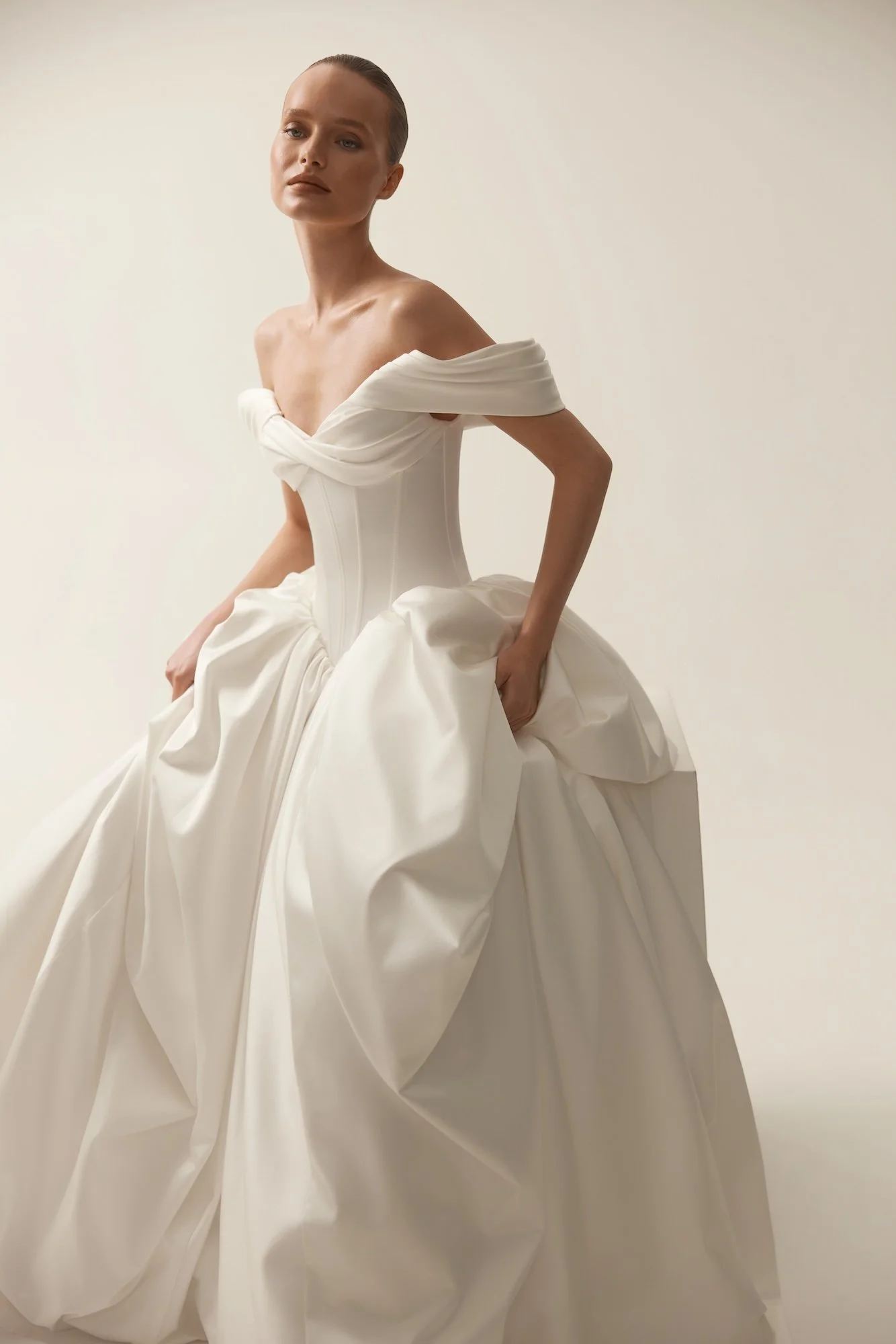 Fashion model wearing an elegant white off-the-shoulder bridal gown by Maison Yana with a corset bodice and flowing skirt.