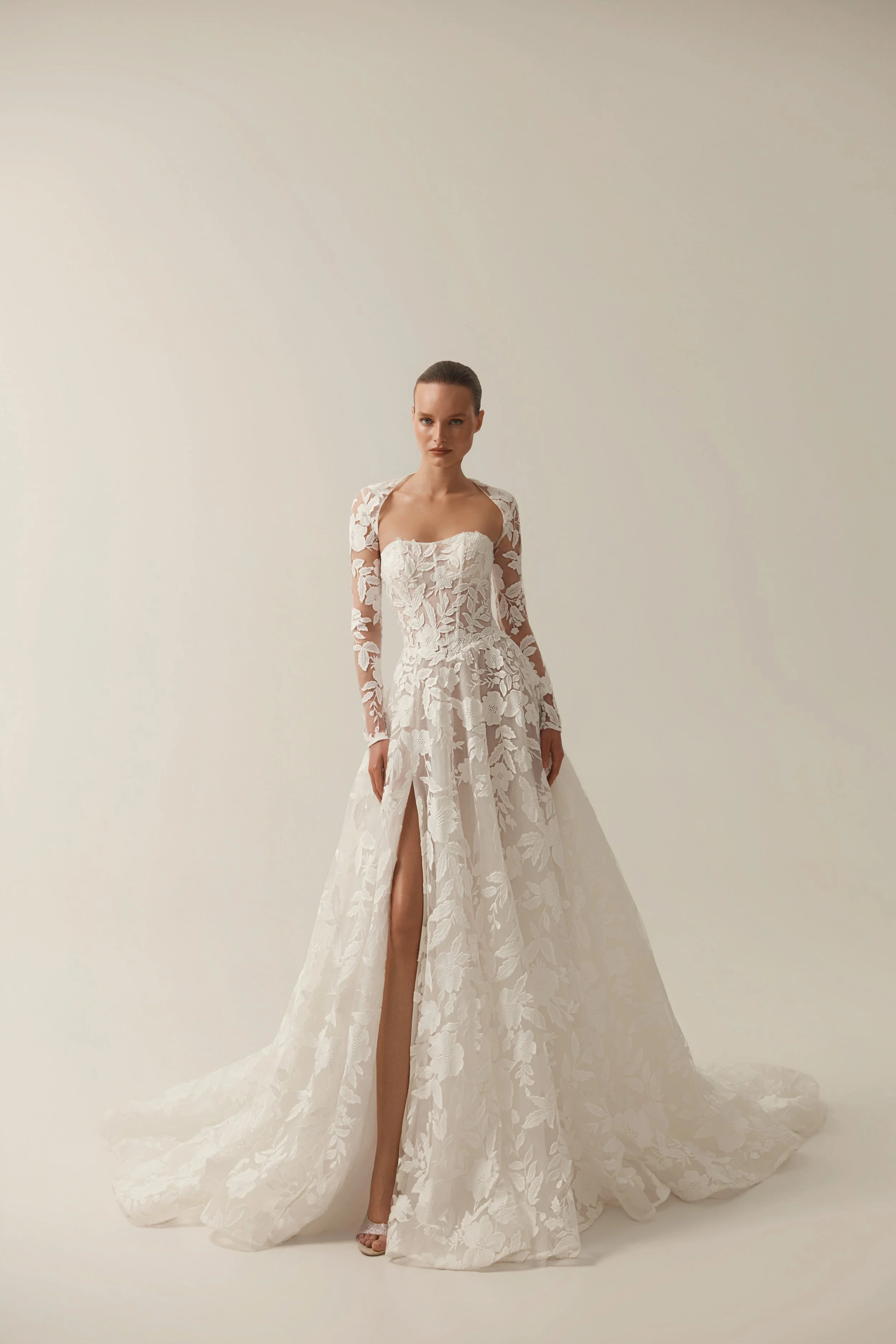Model wearing a white floral lace wedding gown by Maison Yana with a flowing train against a neutral background