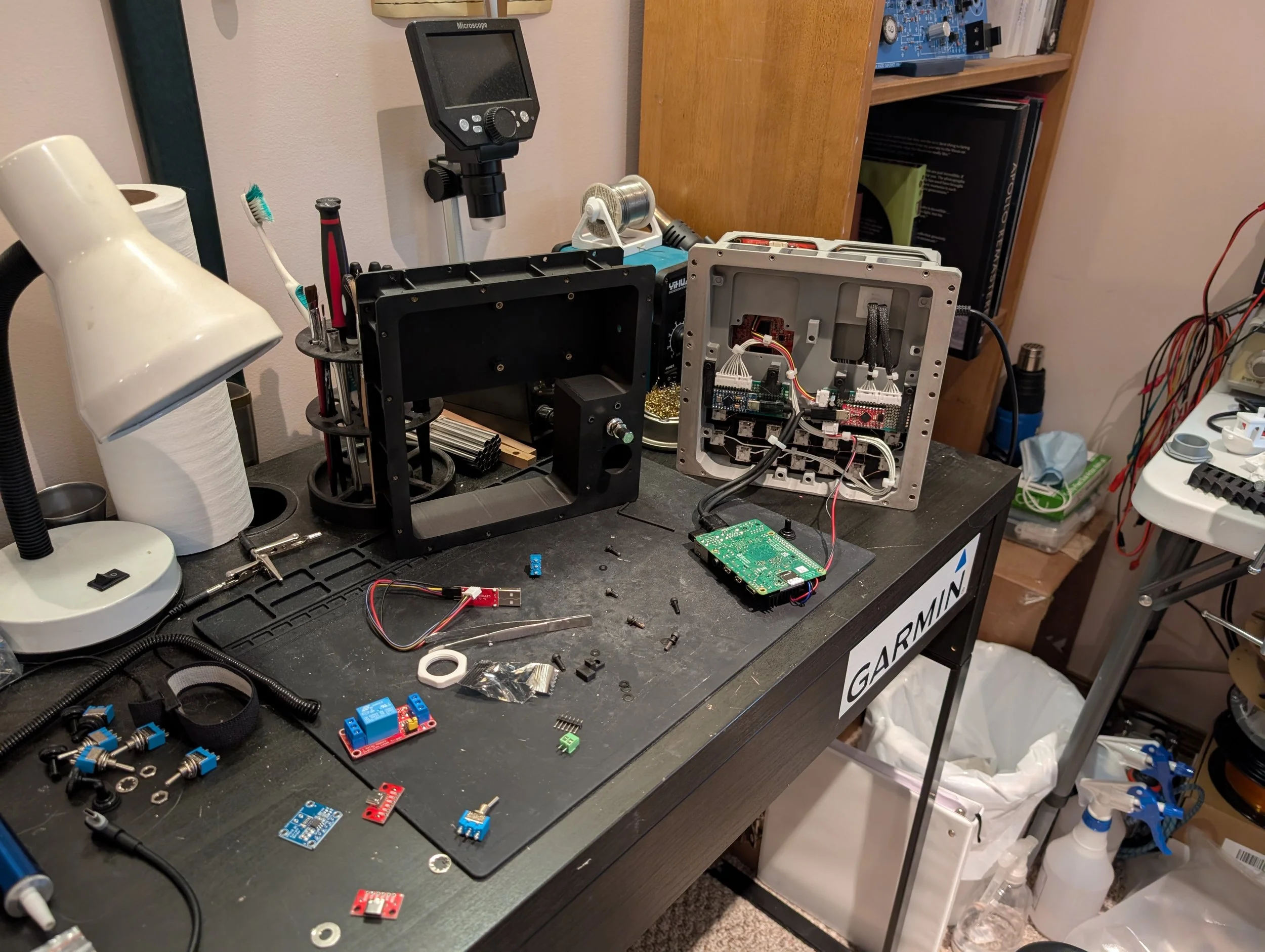 A workbench with various electronic components, tools, wires, circuit boards, and disassembled electronic devices, including a partially opened device revealing internal hardware.