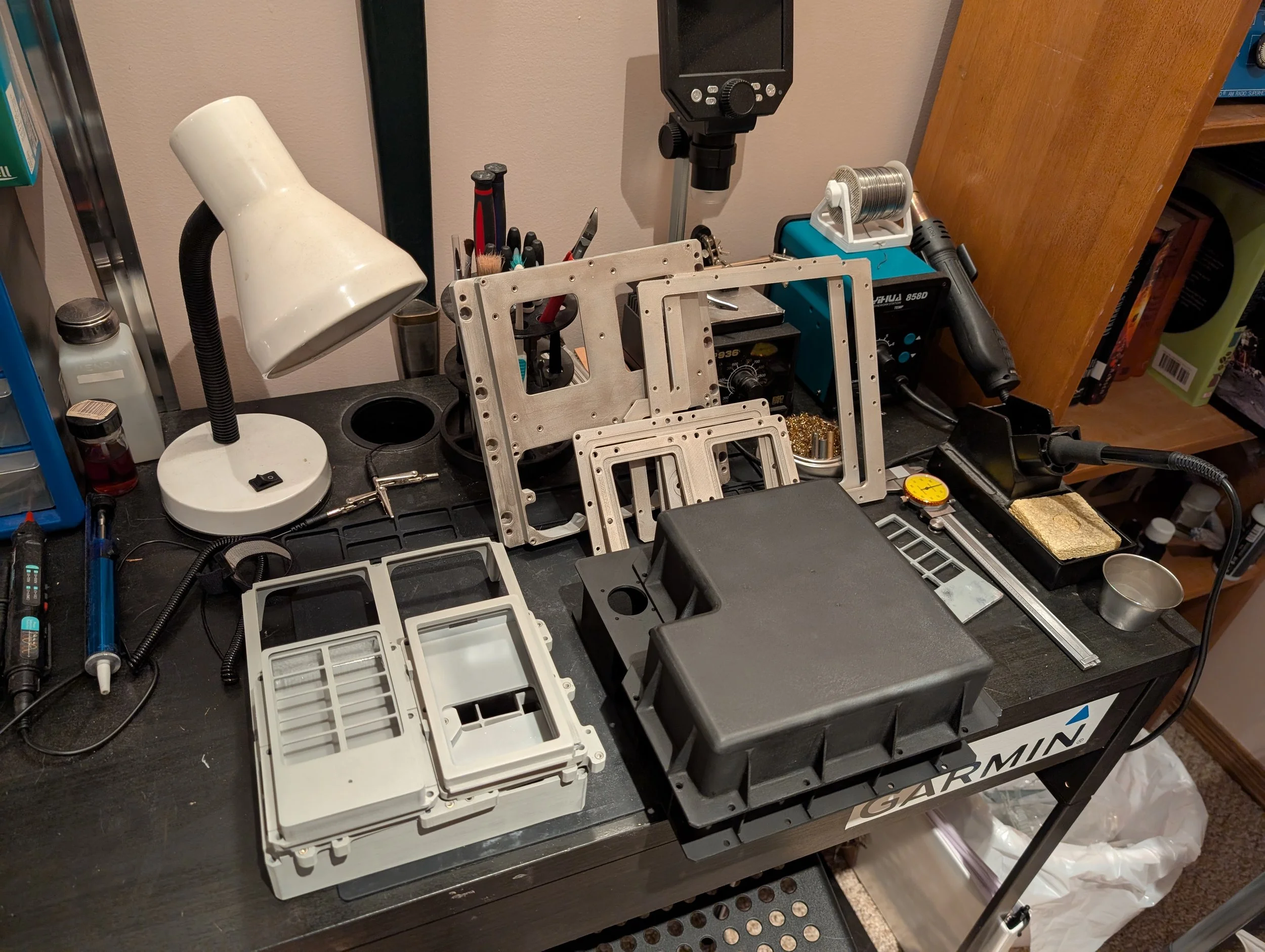 A workbench cluttered with tools and electronic components, including a white desk lamp, a soldering iron, metal frames, a spool of wire, and various screwdrivers and small parts, with cardboard boxes and a wooden shelf in the background.