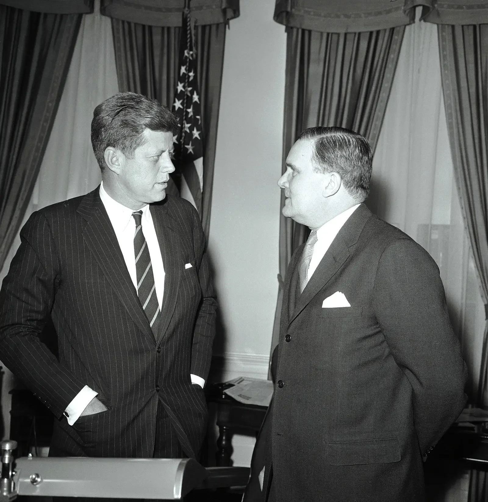 Two men in suits and ties face each other and seem to be having a serious conversation in front of an American flag and curtained windows.